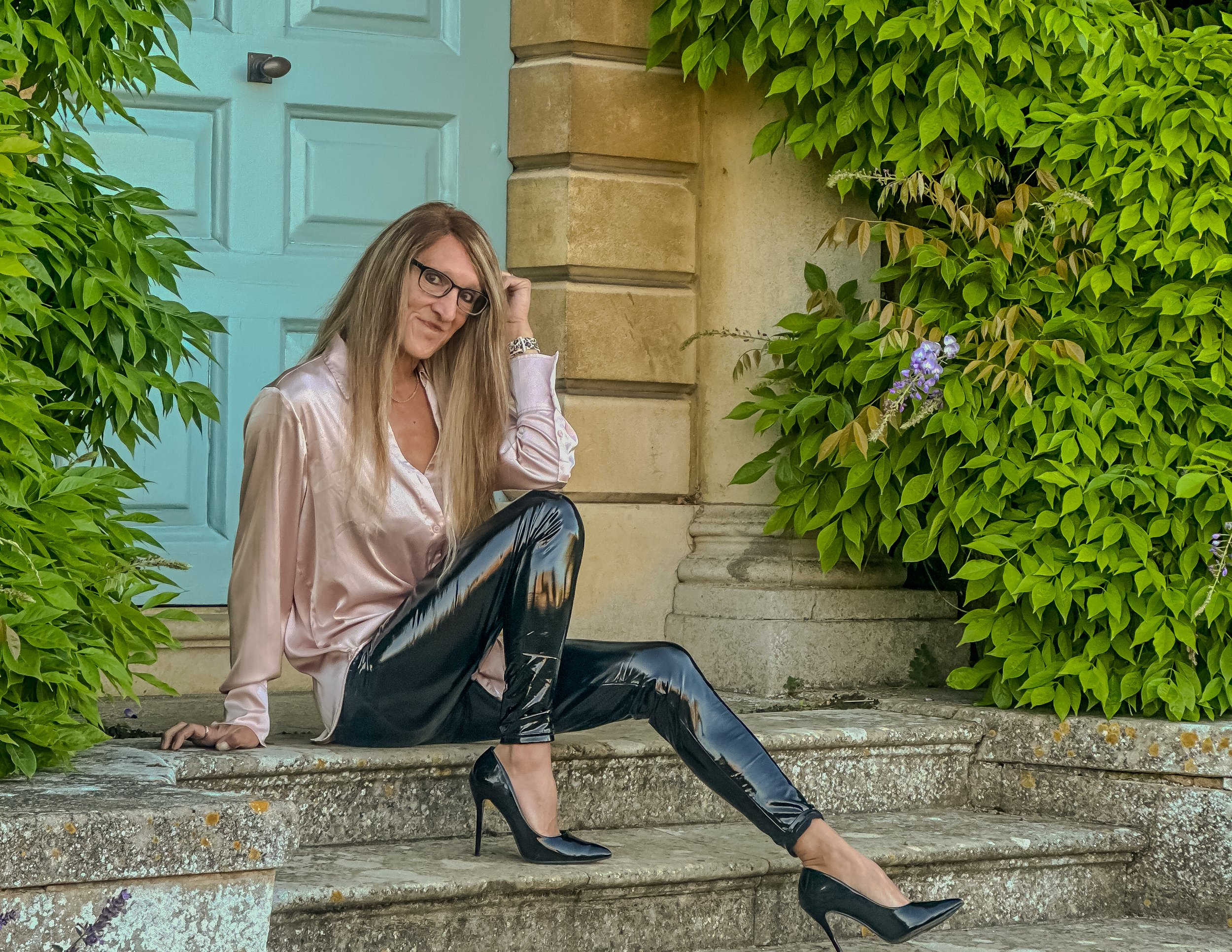 Woman sitting on stone steps with one arm resting on the step behind her, surrounded by lush green foliage, wearing glasses, a light pink silky blouse, black leather pants, and black high heels, with a blue door and a stone building in the background