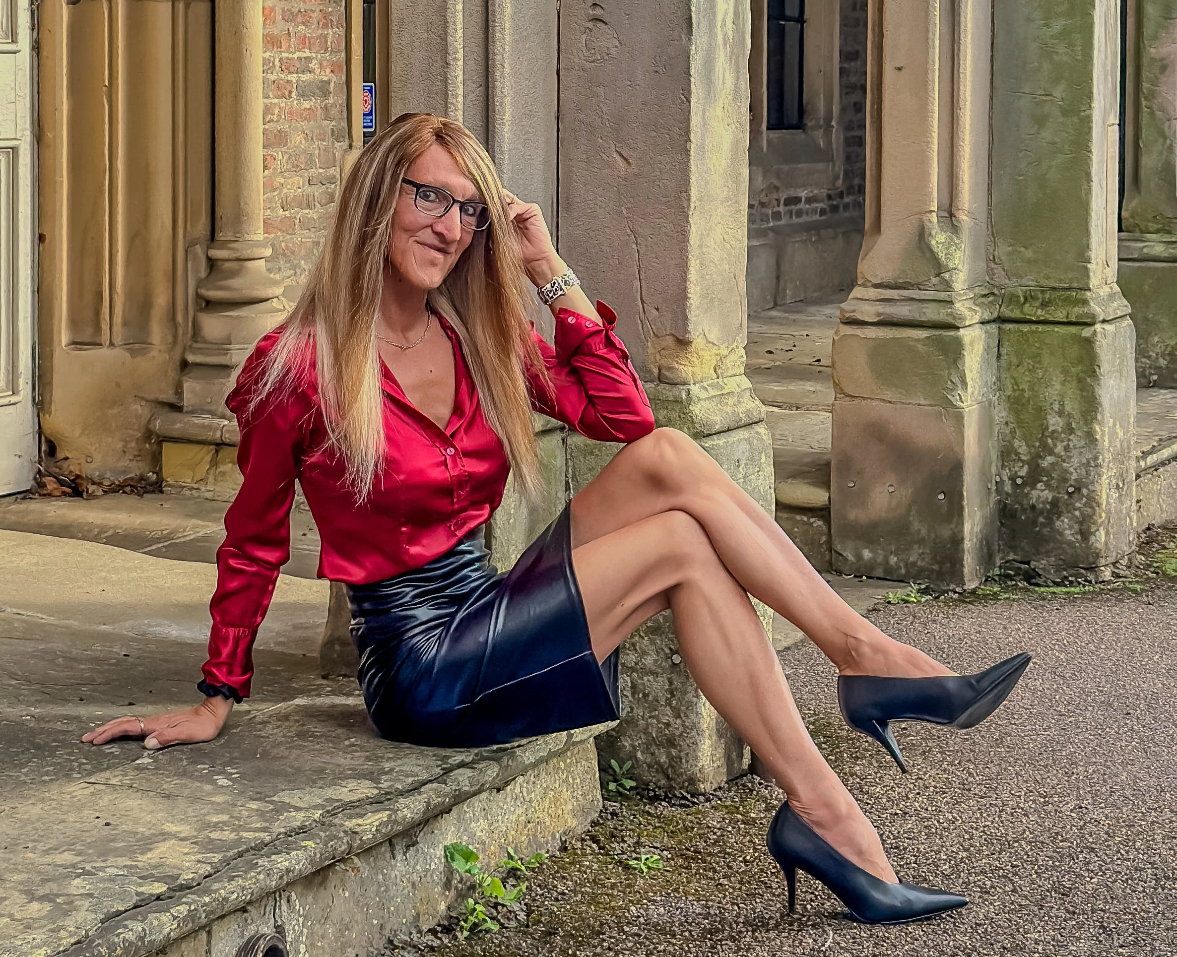 A woman with long blonde hair, glasses, and a red satin blouse sitting on stone steps near a brick building with columns, posing for the camera with her legs crossed and hand near her head.