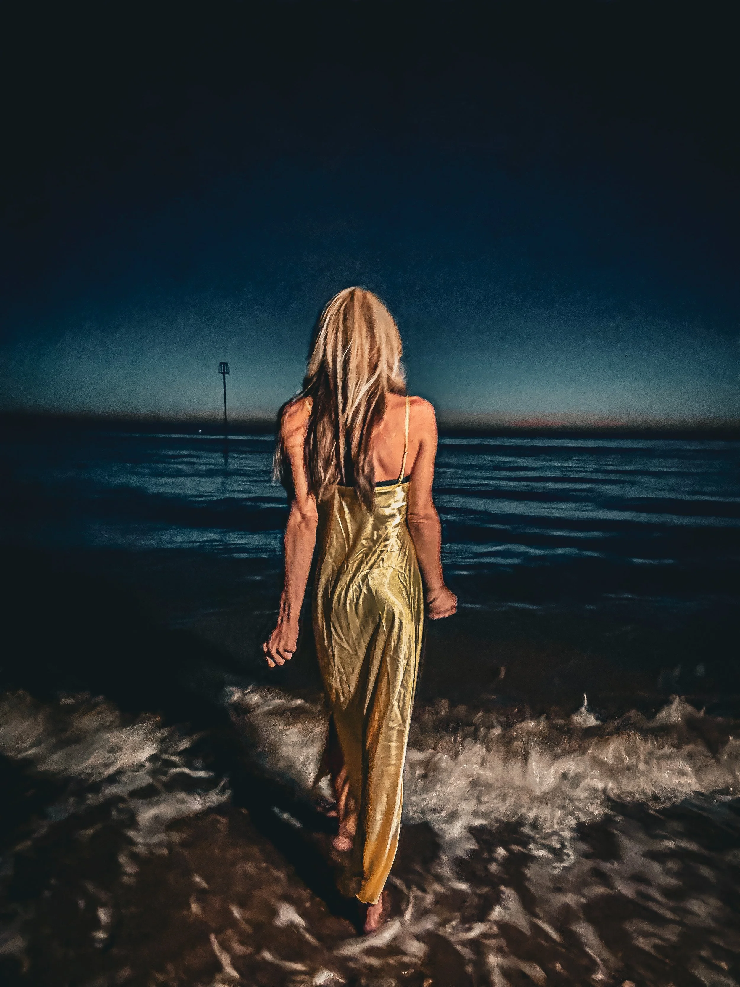A woman with long blonde hair wearing a yellow dress standing in the surf at night, looking at the ocean horizon.