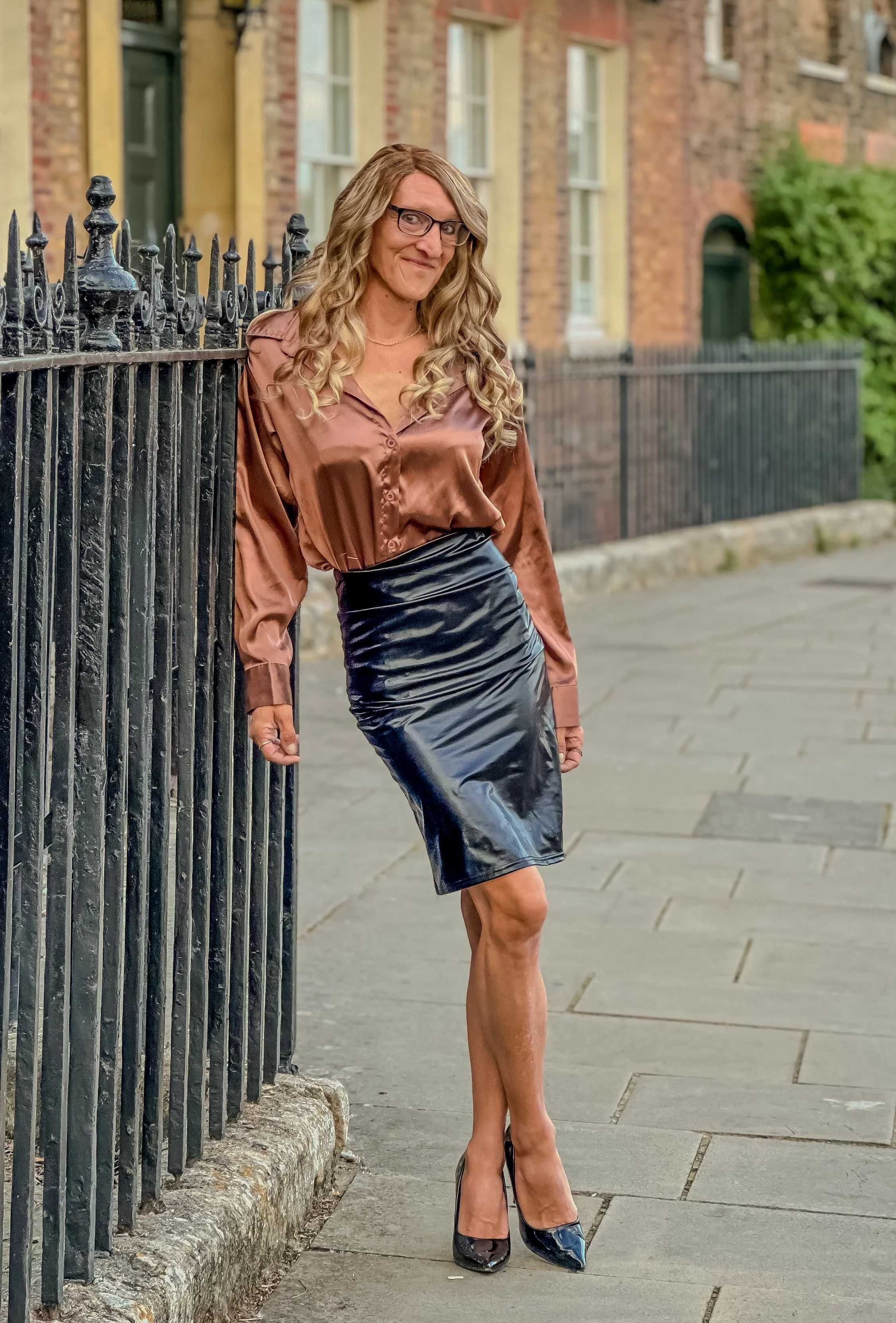 A woman with long curly blonde hair, glasses, wearing a shiny brown blouse, a shiny black pencil skirt, and black high heels, leaning against a black iron fence on a sidewalk in an urban neighborhood.