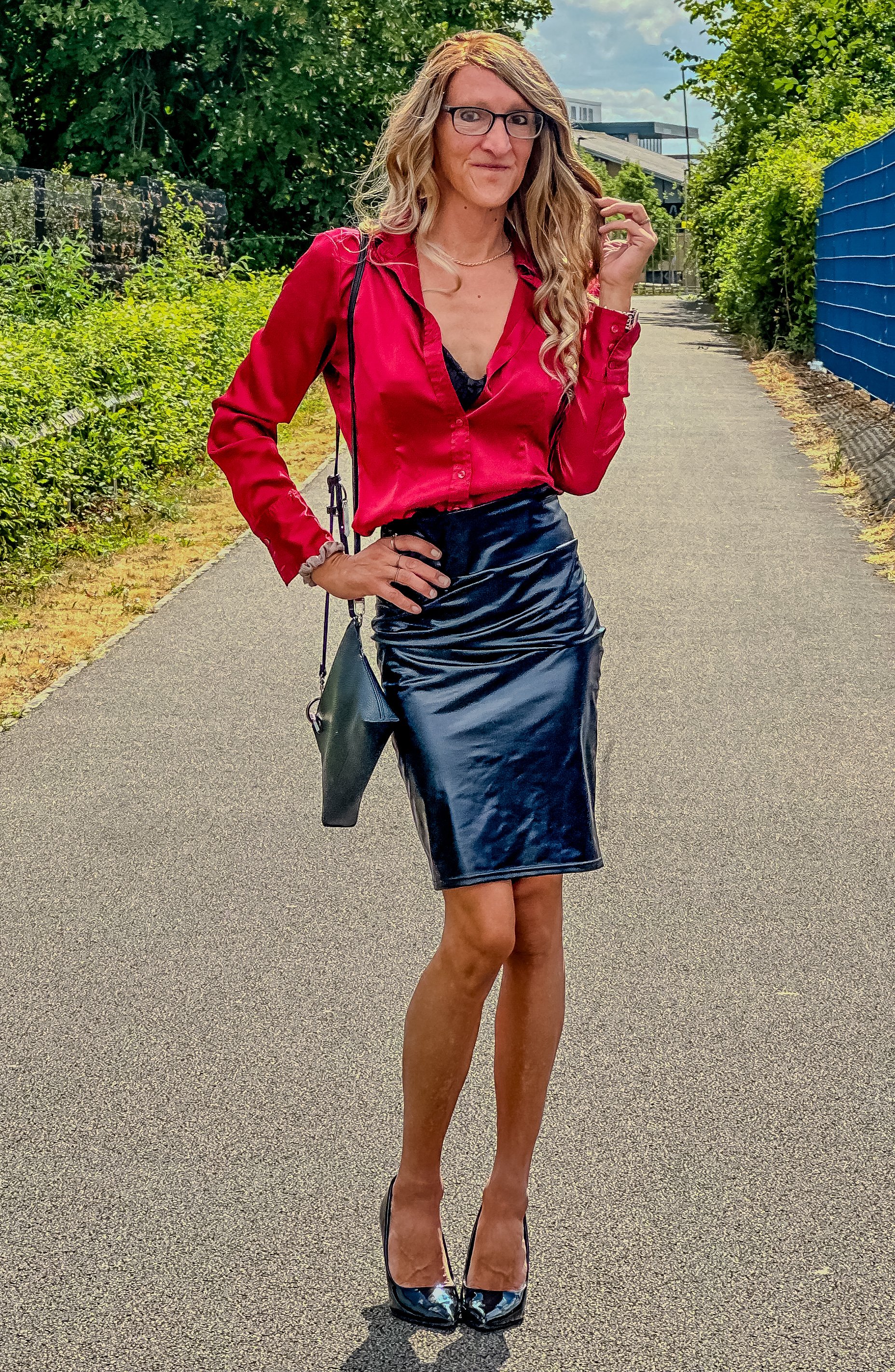 A woman wearing a red satin shirt, a black leather skirt, high heels, glasses, and a necklace, standing outdoors on a sidewalk with greenery on both sides.