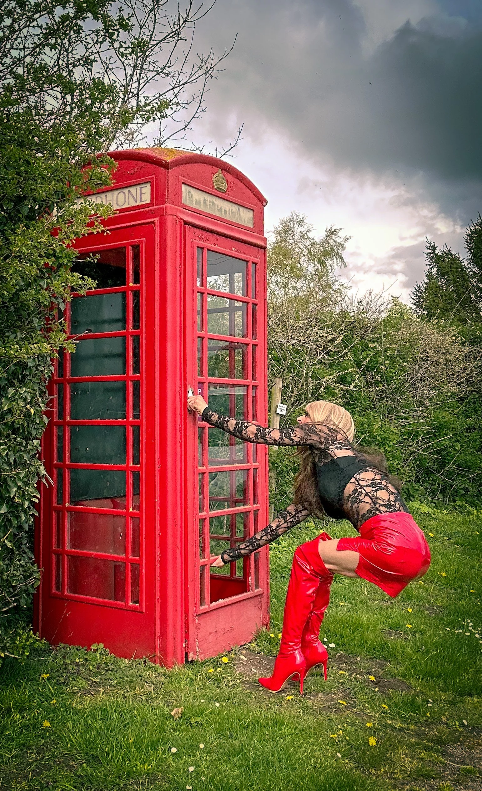 A woman with long hair, wearing a black lace top, red leather pants, and red high-heeled boots, crouches near a red British telephone booth outdoors, with trees and cloudy sky in the background.