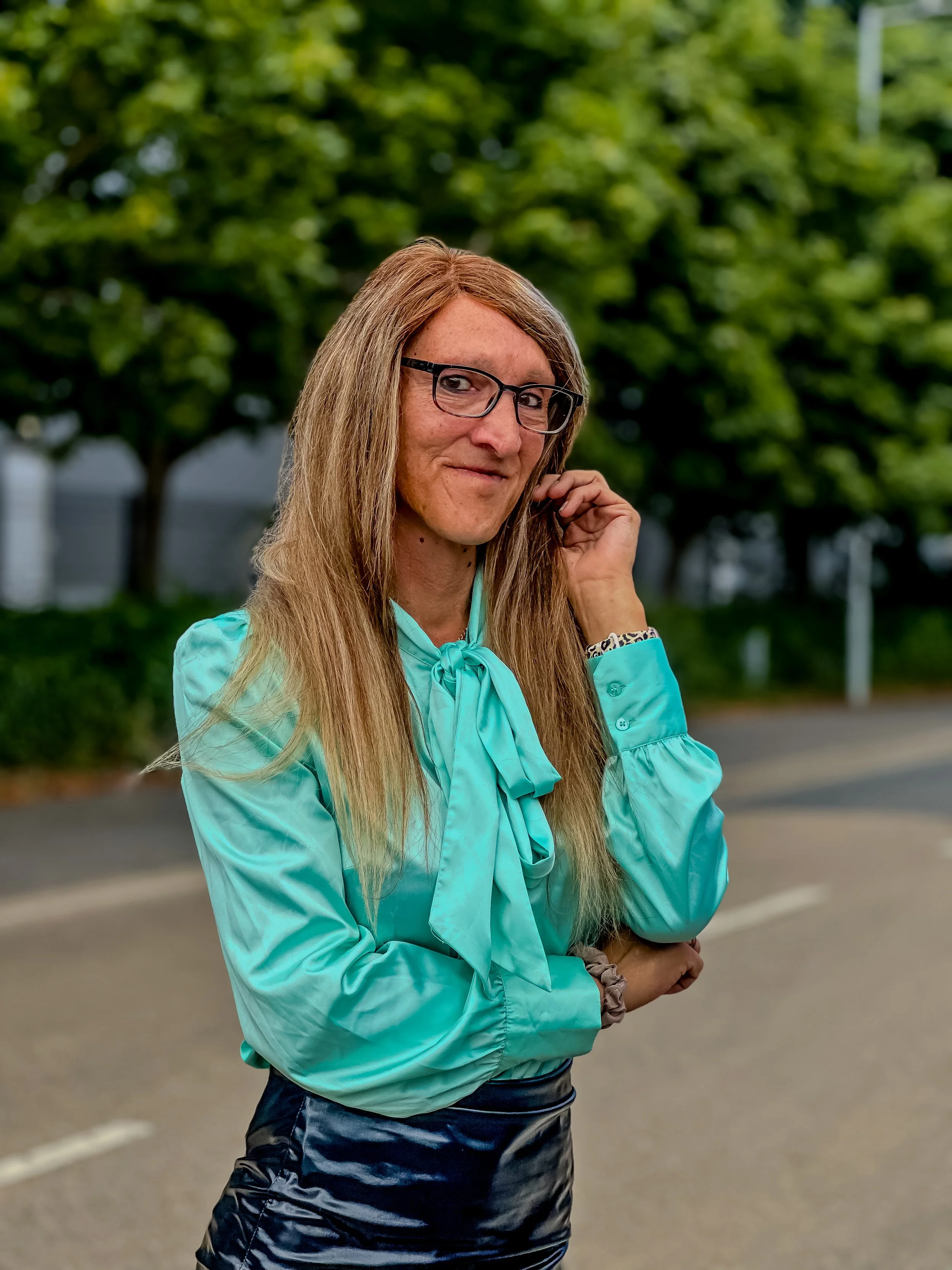 A woman with long blonde hair, glasses, wearing a turquoise blouse and a black shiny skirt, stands outdoors near a road with green trees in the background.