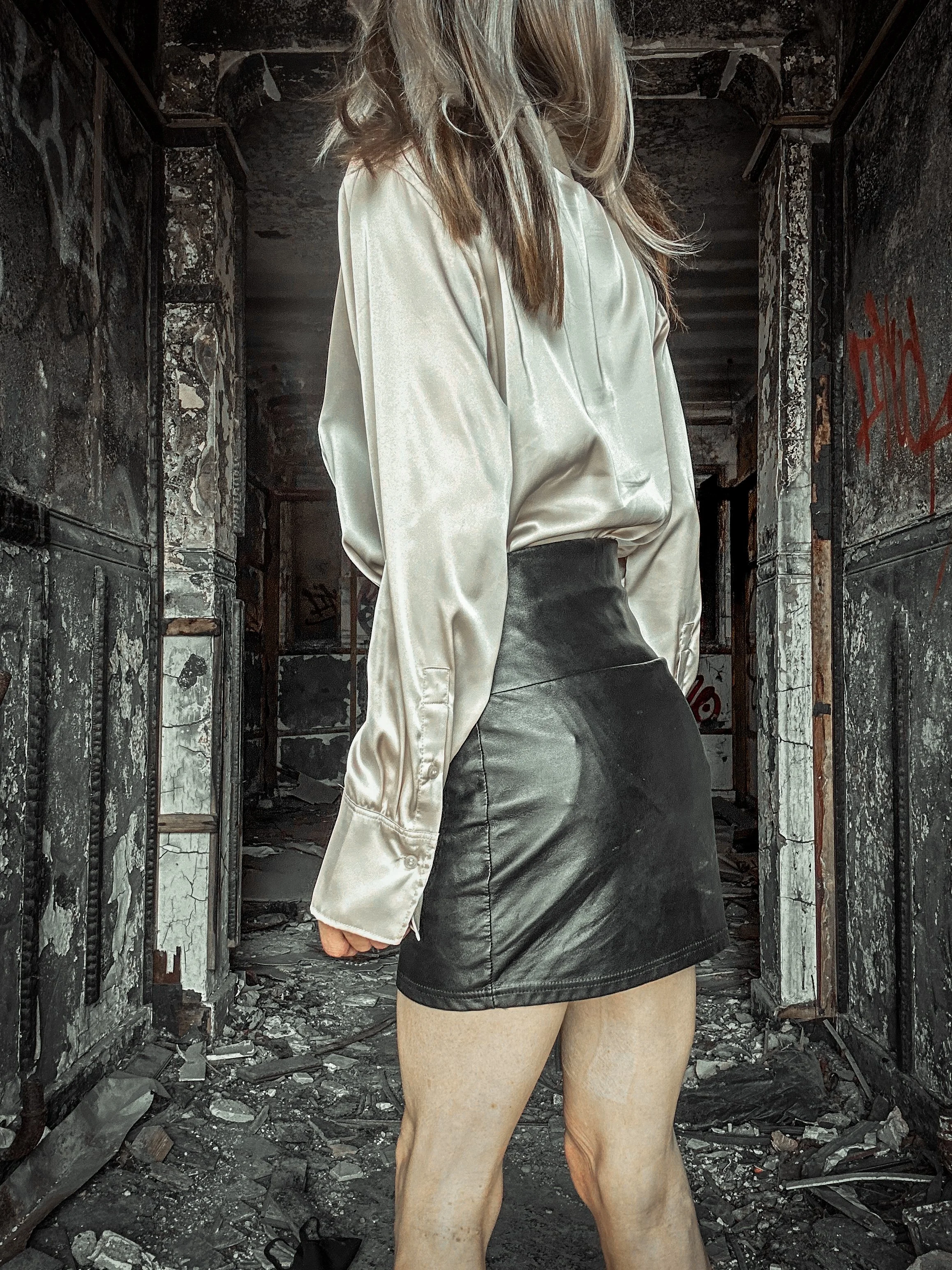 A woman with long hair wearing a satin blouse and a leather skirt, standing in a ruined building with debris on the ground and graffiti on the walls.