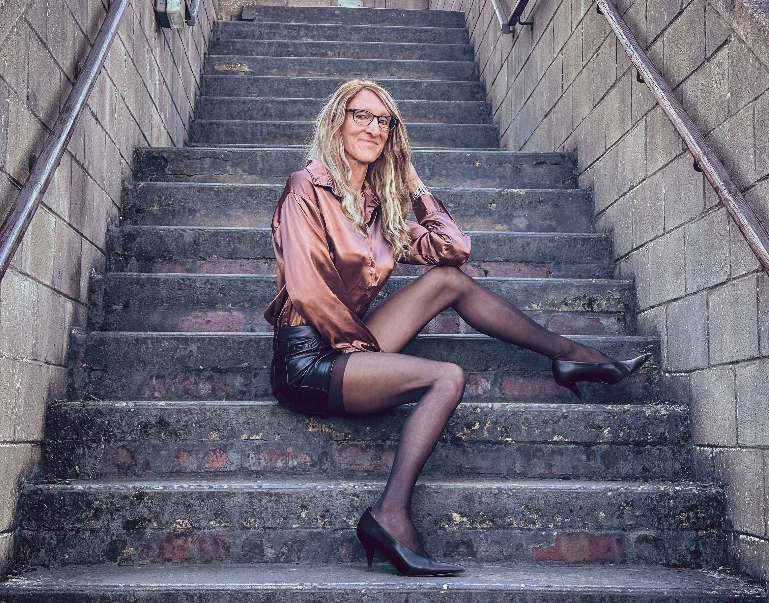 A woman with long blonde hair, glasses, and a satin pink blouse sitting on outdoor concrete steps, wearing black heels and stockings.