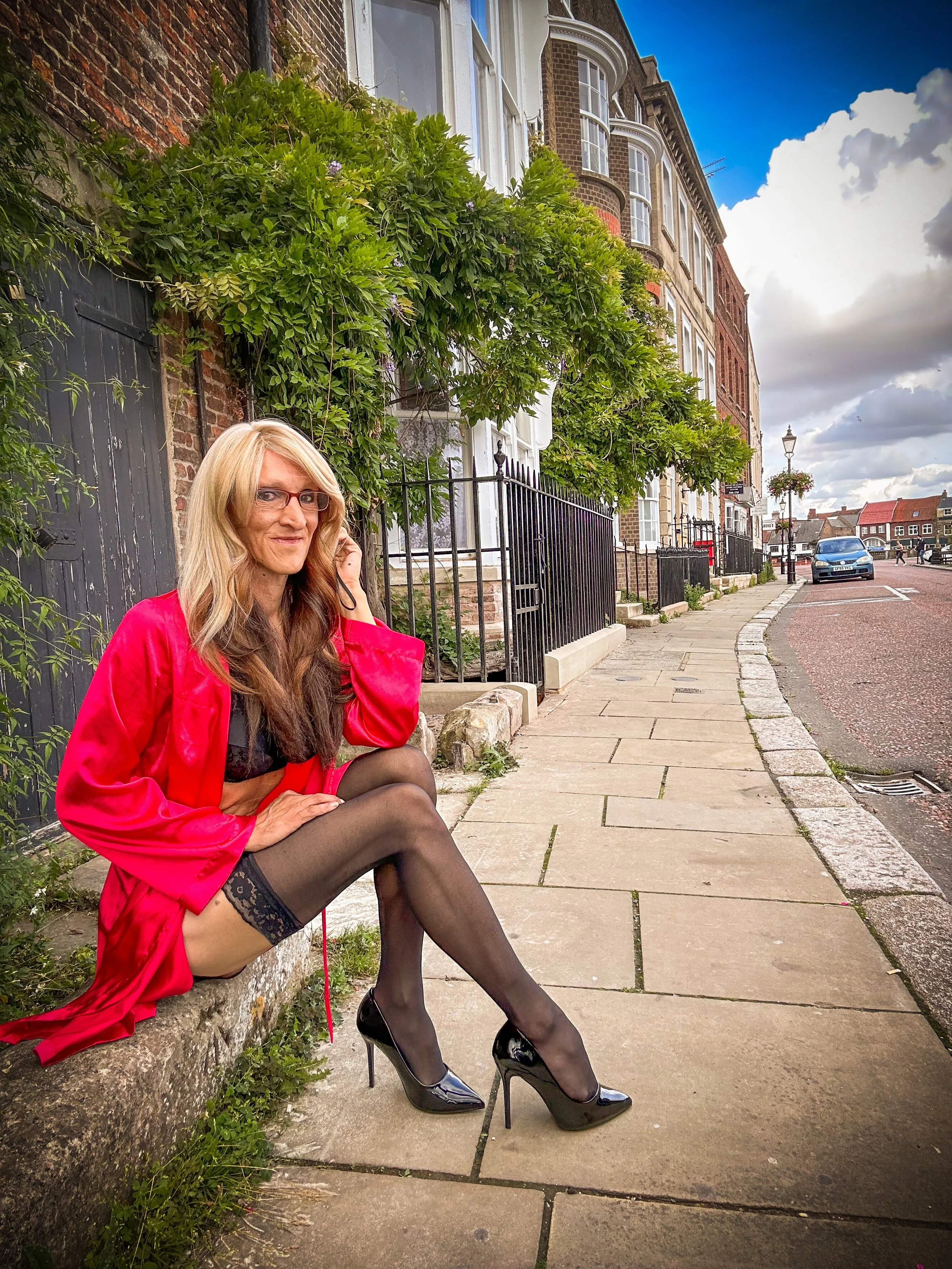 A woman wearing black stockings and high heels, sitting on a sidewalk in front of a low stone wall and green foliage. She is dressed in a red satin robe and black lace lingerie, with long wavy hair and glasses, talking on a cell phone. The background
