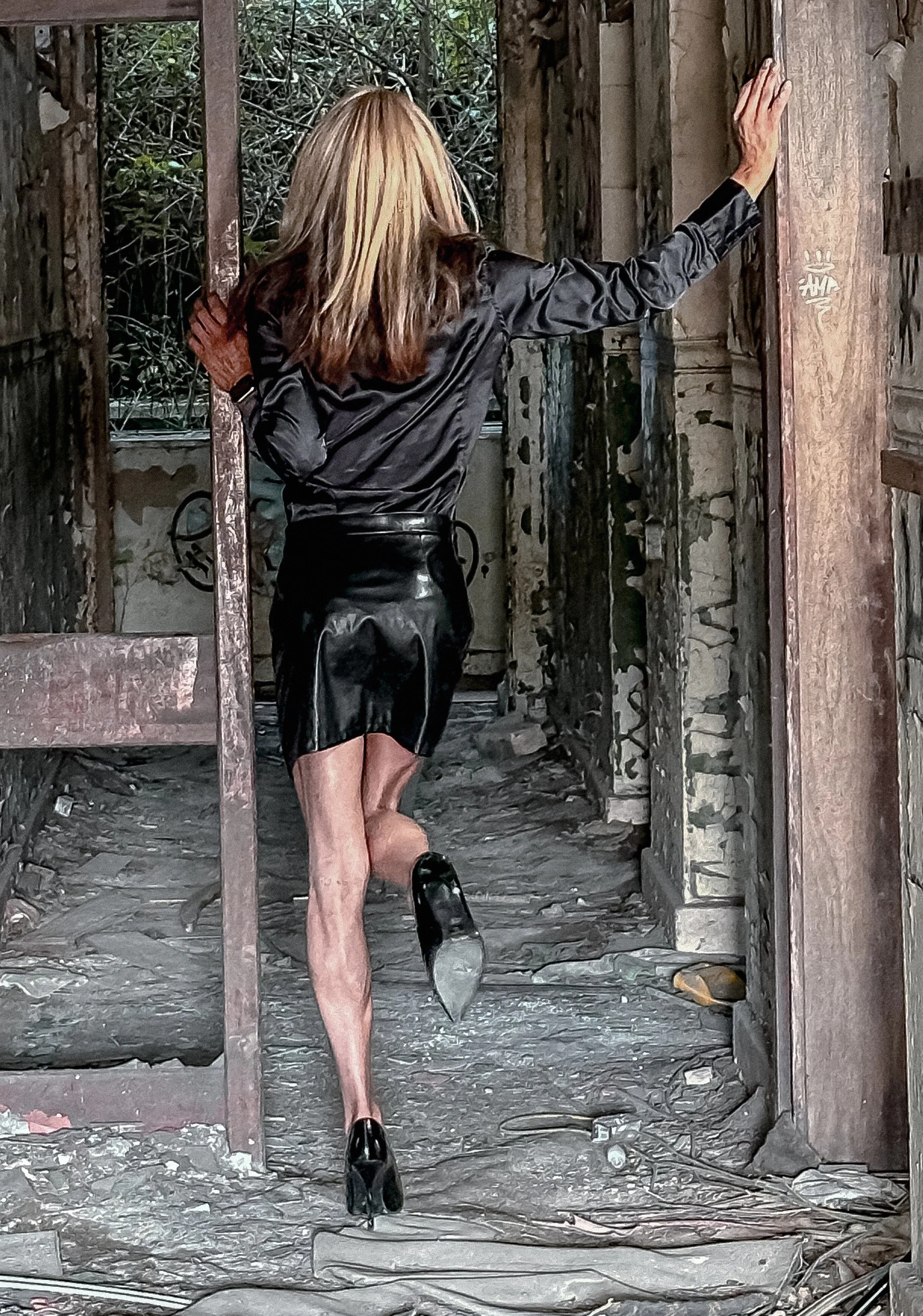 A woman with blonde hair dressed in a black leather jacket and skirt, wearing high heels, is seen walking away in an abandoned, dilapidated building with graffiti on the walls. The scene appears to be outdoors with overgrown vegetation visible throug