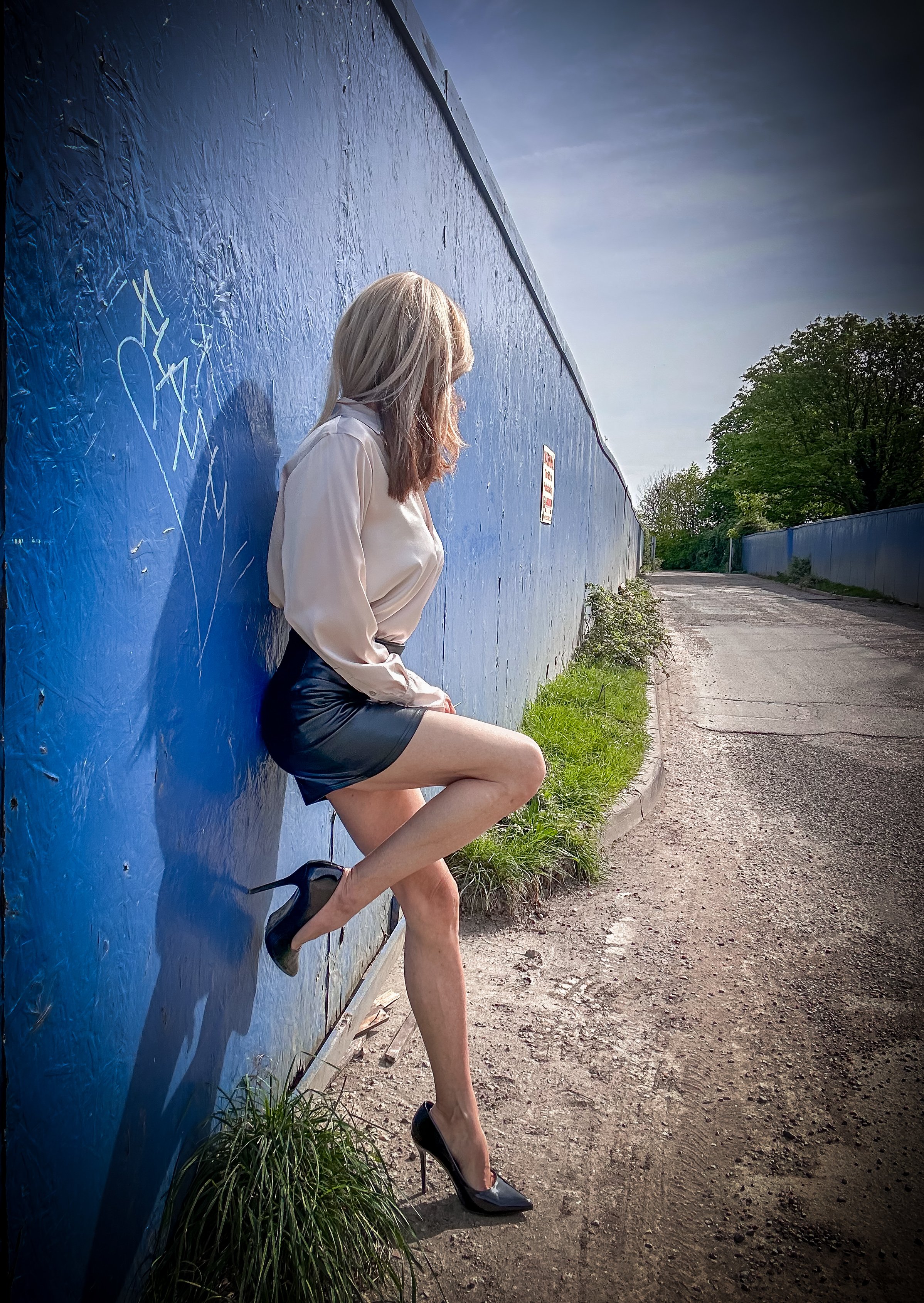 A woman with blonde hair, wearing a beige blouse, black leather skirt, and black high heels, is leaning against a blue wall with graffiti. She is standing on a dirt path with green grass on the sides and trees in the distance.