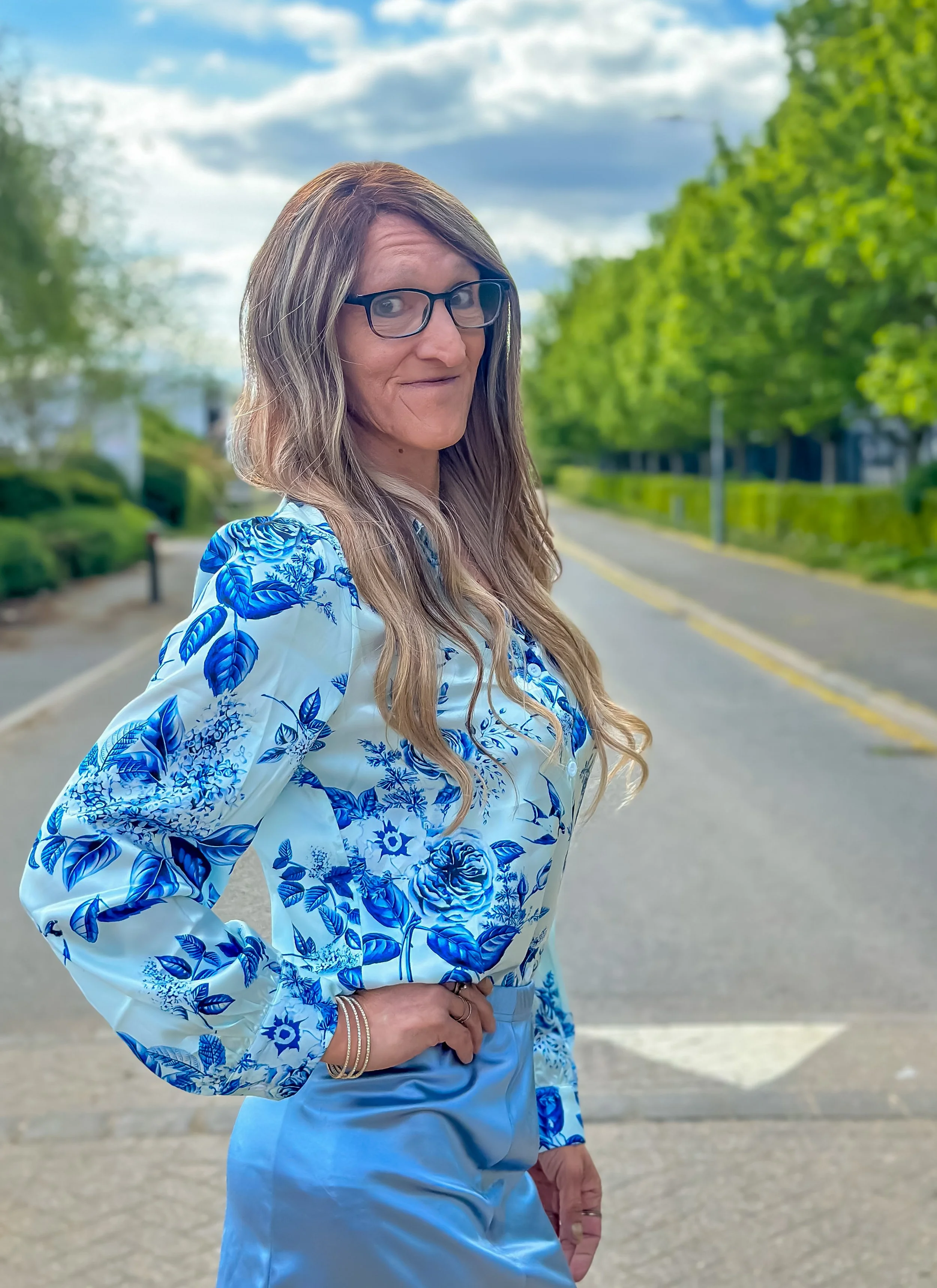 A woman with long, wavy hair wearing glasses and a blue-and-white floral blouse standing outdoors on a sidewalk with trees and a cloudy sky in the background.