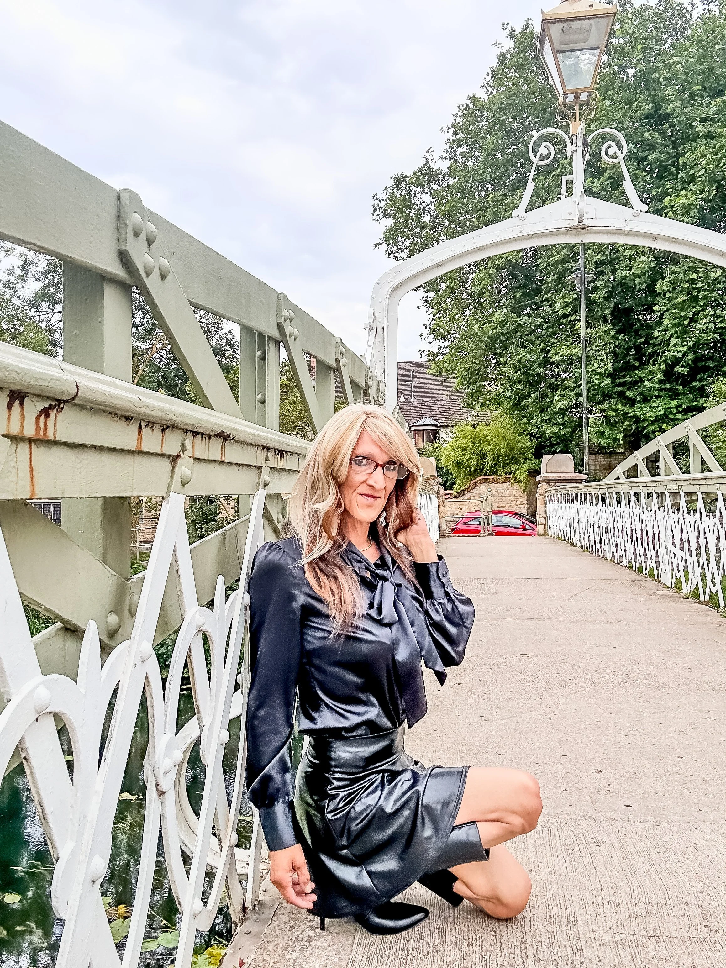 Woman with blonde hair and glasses sitting on the sidewalk next to a white decorative metal bridge, wearing a black leather jacket, a black satin skirt, and black boots, in an outdoor setting with a tree and houses in the background.