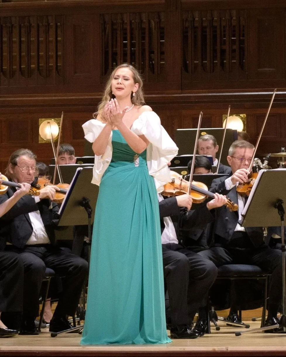 A woman in a turquoise gown performing in front of an orchestra on stage, with some of the orchestra members playing violins behind her.