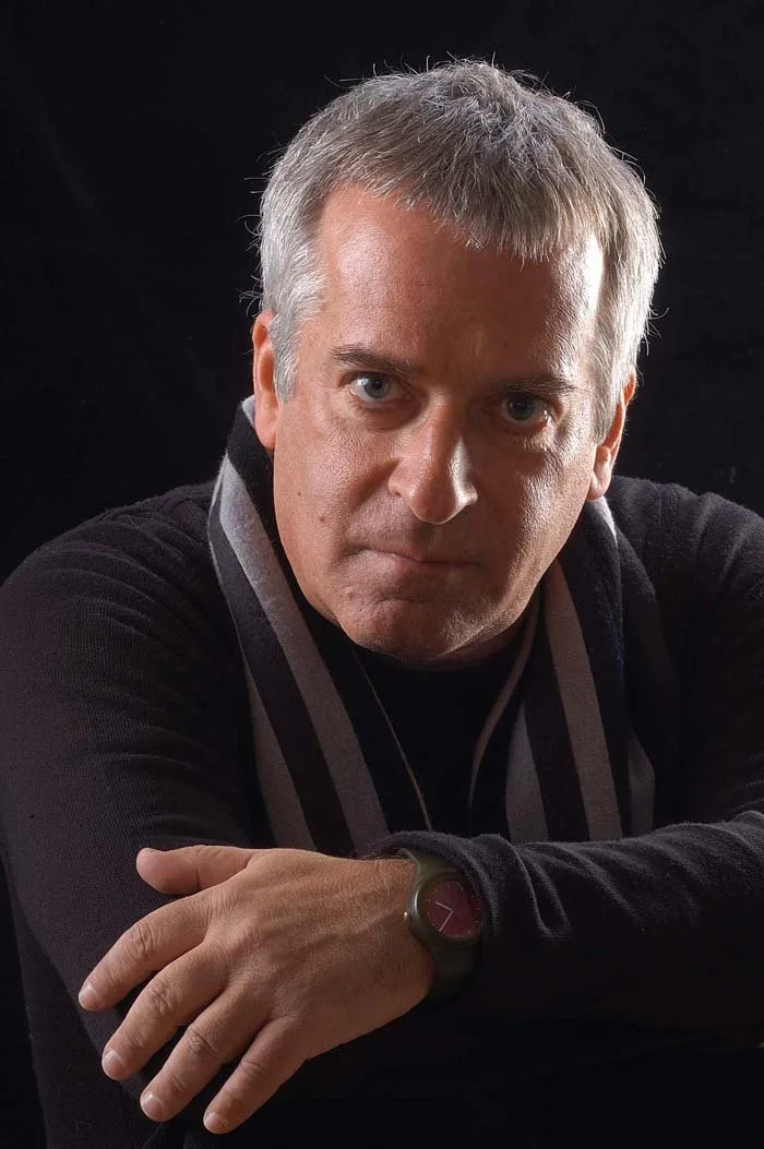 A middle-aged man with short gray hair wearing a black shirt and a dark striped scarf, looking directly at the camera with a serious expression, against a black background.