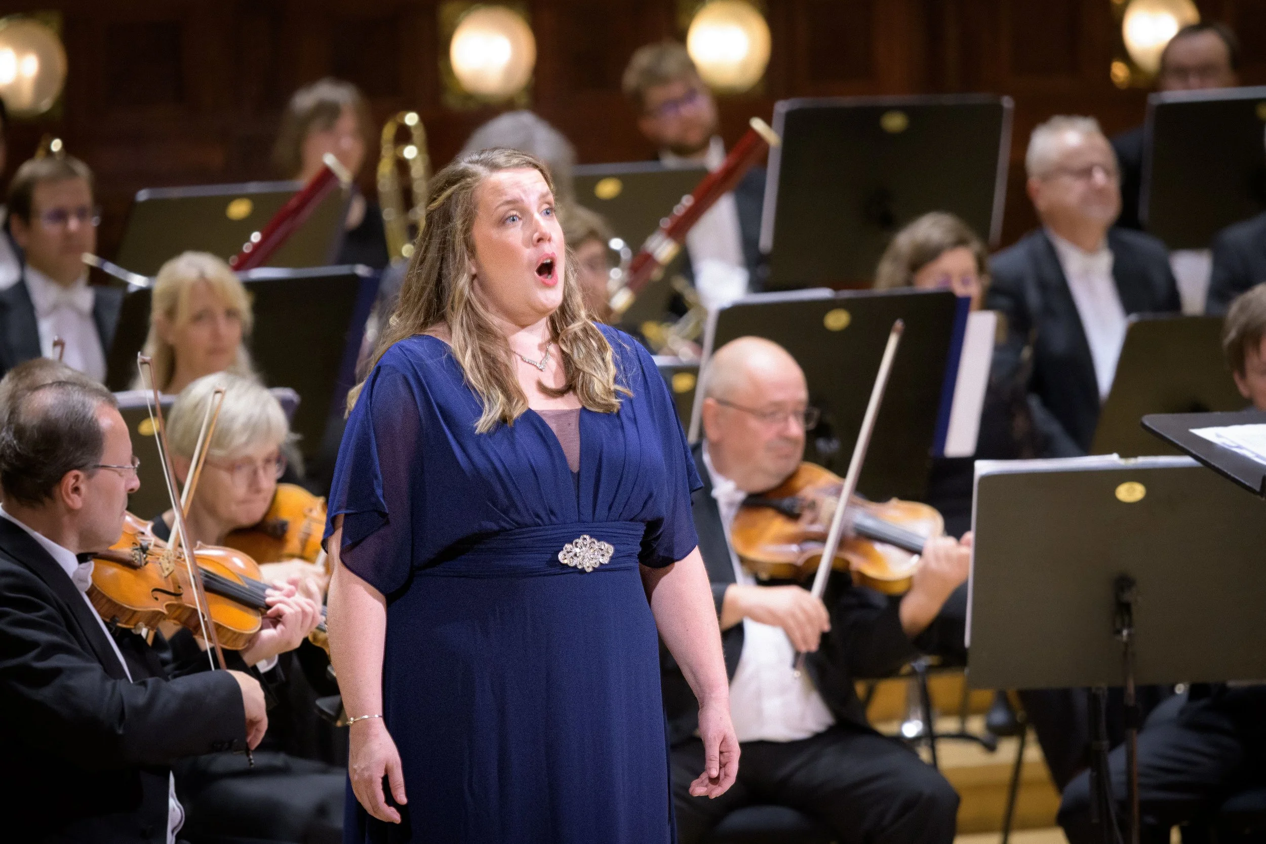 A woman in a blue dress singing during a classical orchestra performance with musicians playing violins and cellos behind her.