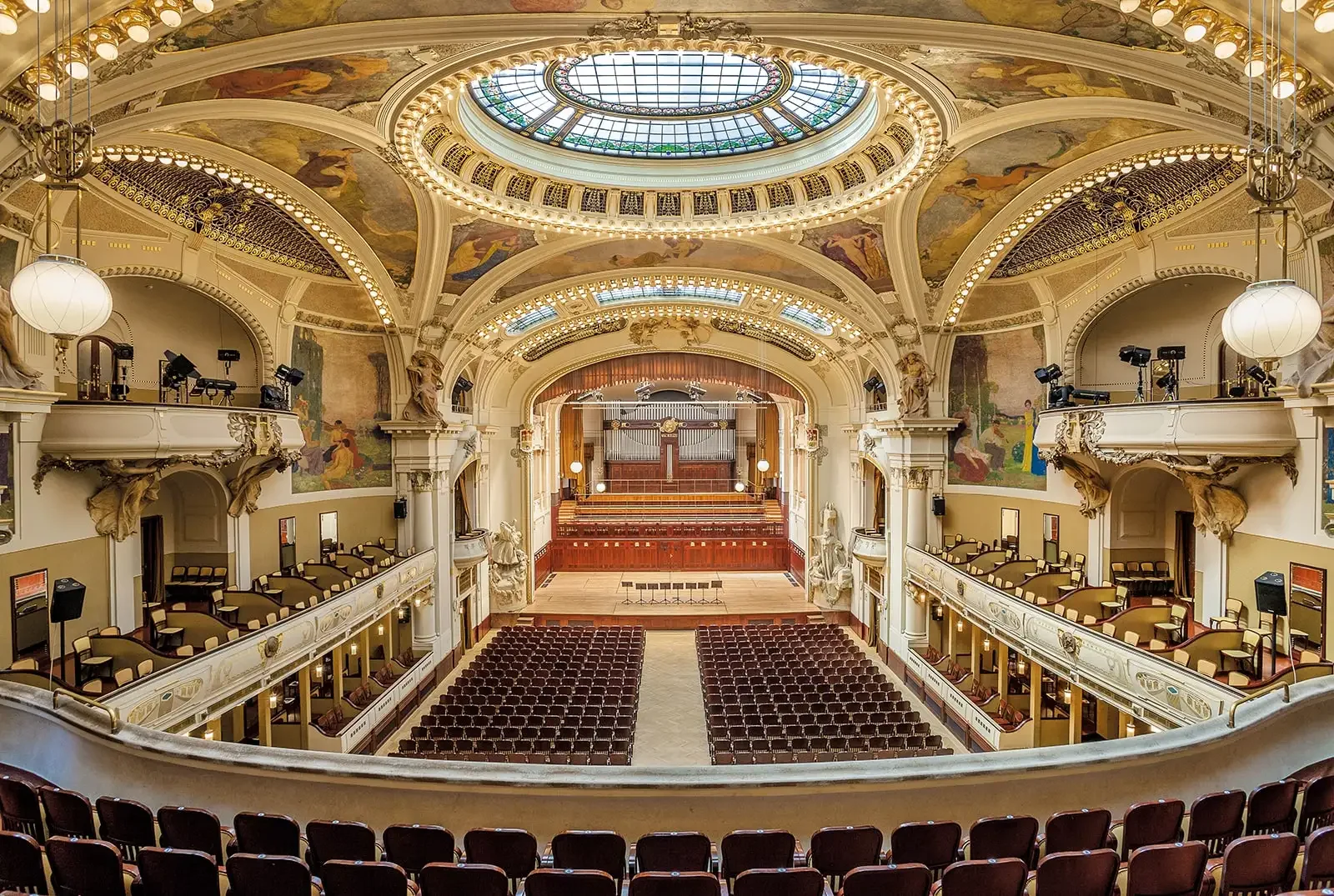 Interior of a grand, historic theater with ornate architecture, a large stained glass skylight, multiple balconies, and a stage at the front.