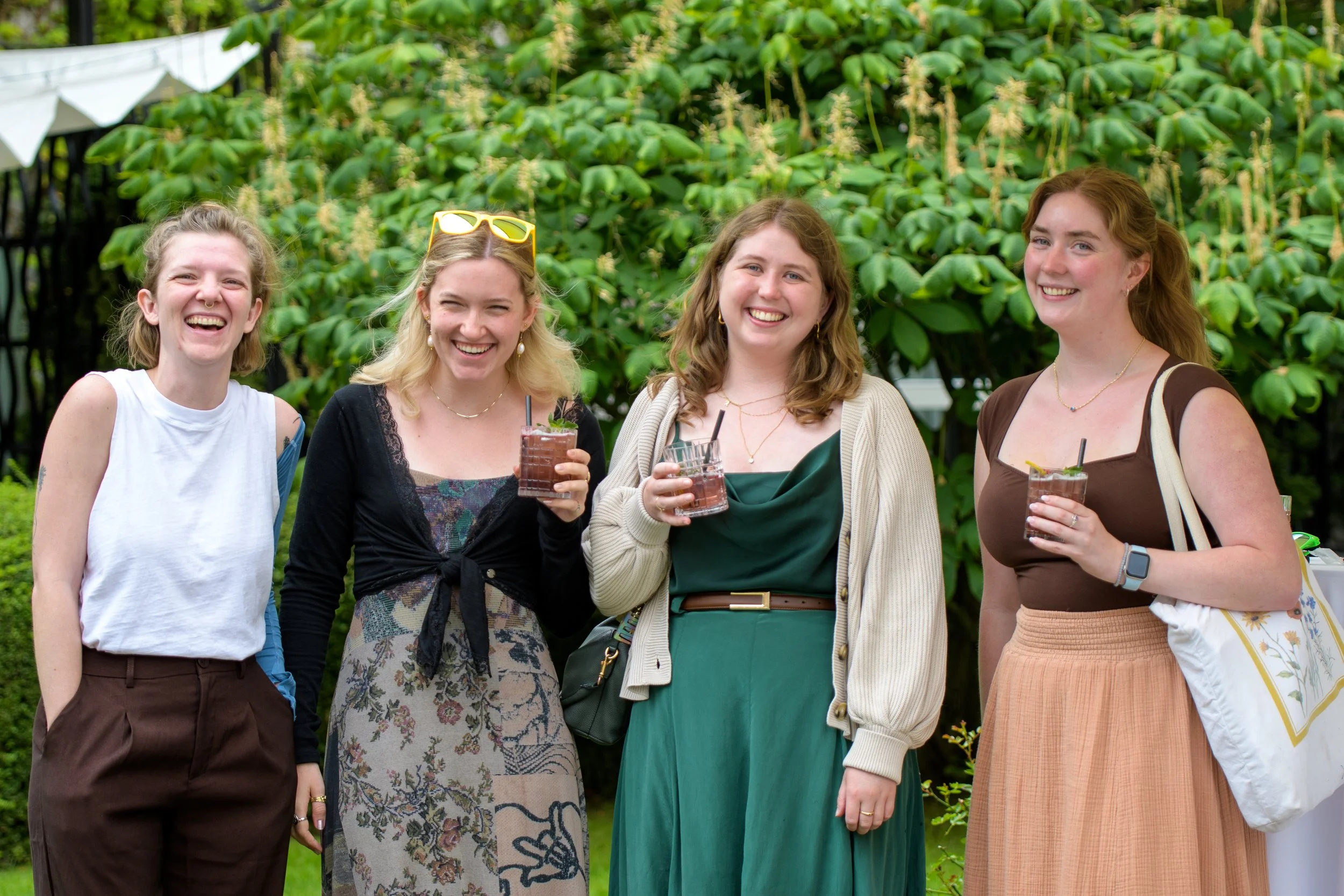 Four women standing outdoors in front of leafy green bushes, smiling and holding glasses of drinks, enjoying a social gathering.