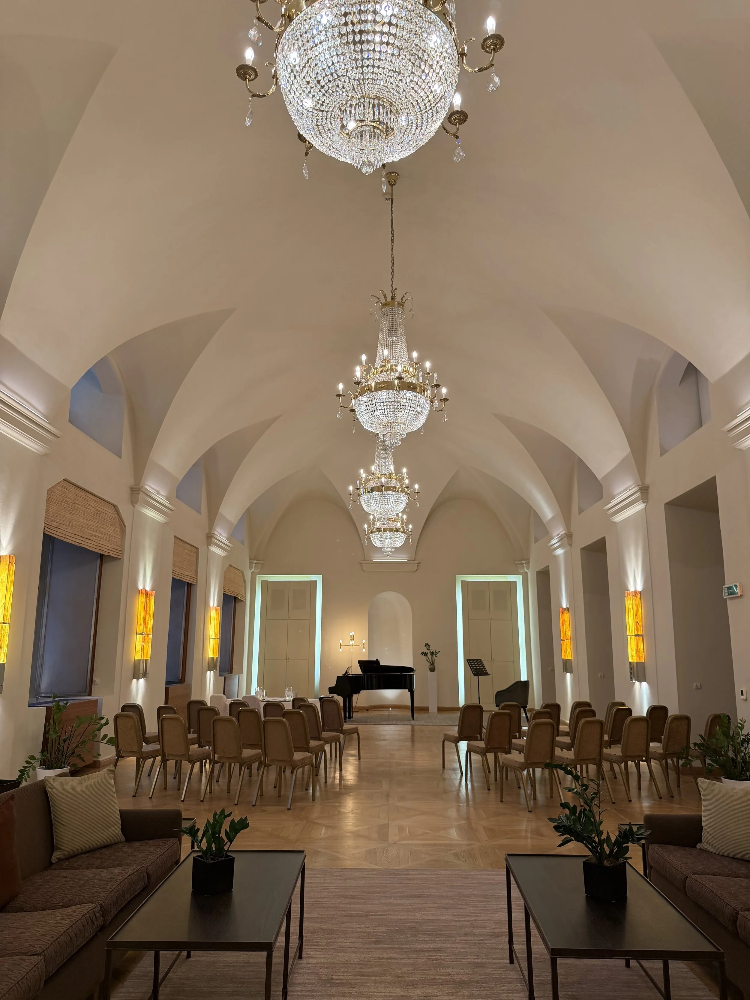 Elegant room with high vaulted ceilings, multiple sparkling chandeliers, rows of chairs, a grand piano, and soft lighting, suggesting a setting for a performance or formal event.