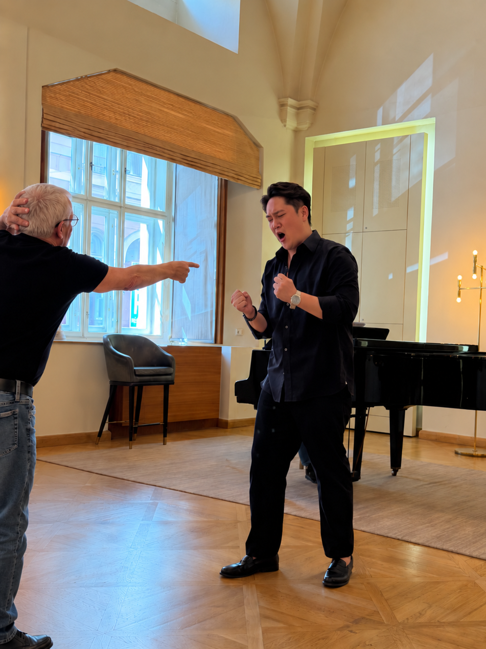 A young man in black clothing with a watch on his left wrist is singing passionately with his eyes closed, fists clenched, and standing in a room with natural light, near a grand piano and an older man pointing at him in anger or frustration.