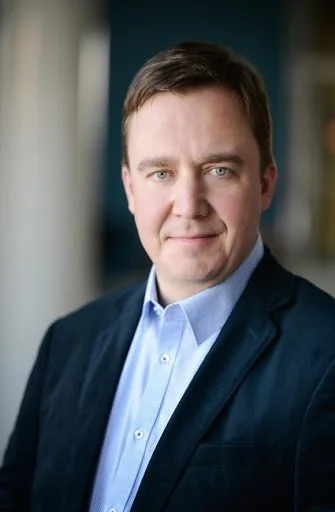 Professional headshot of a man in a dark suit and light blue shirt, smiling slightly, in an indoor setting with a blurred background.