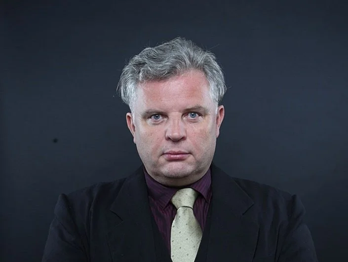 A man with gray hair and blue eyes wearing a black suit, maroon shirt, and beige tie, looking directly at the camera against a dark background.