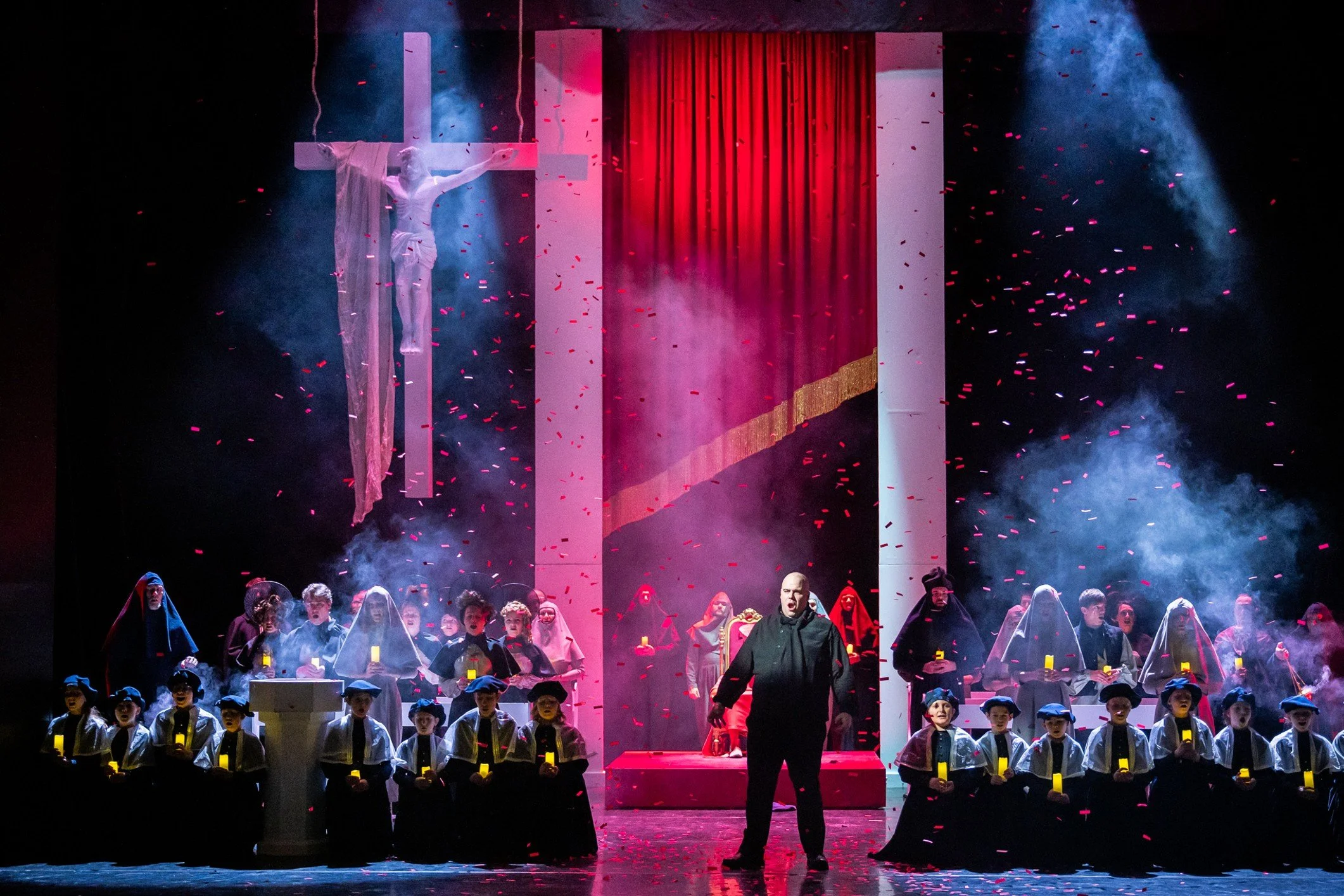 A theatrical performance featuring a large group of children dressed in costumes, standing on stage in front of a red curtain, with dramatic lighting, smoke effects, and red and pink confetti falling from above. A figure in black stands at center stage.