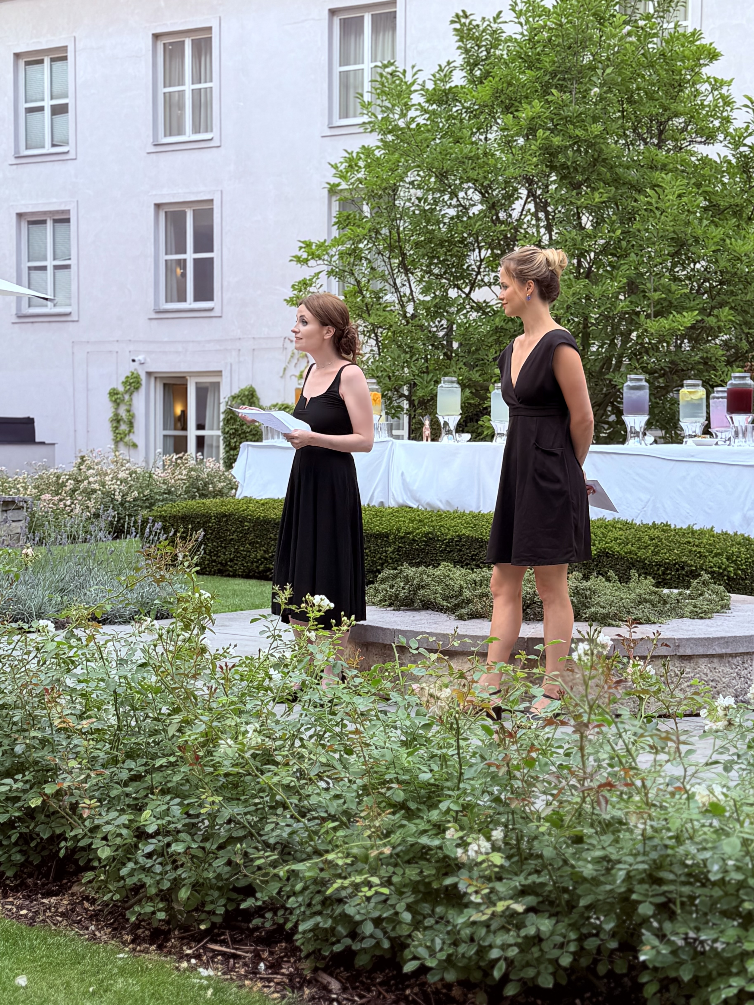 Two women in black dresses standing outdoors in a garden area with a table of colorful drinks behind them. One woman is holding papers and reading, the other stands with her hands behind her back. There is a large green tree and a white building with multiple windows in the background.