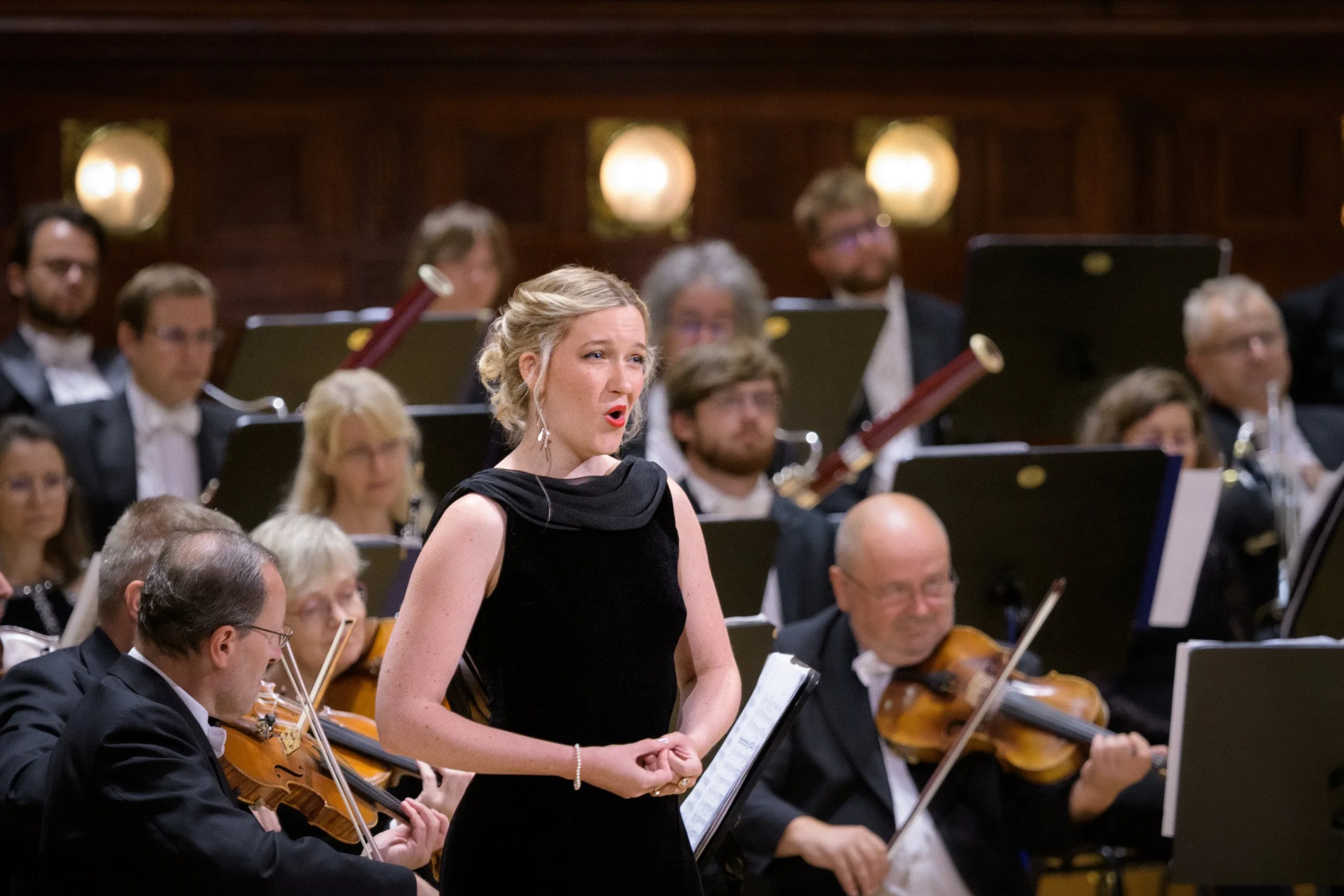 A woman in a black dress singing during an orchestra performance, with multiple orchestra members playing string instruments in the background in a concert hall.