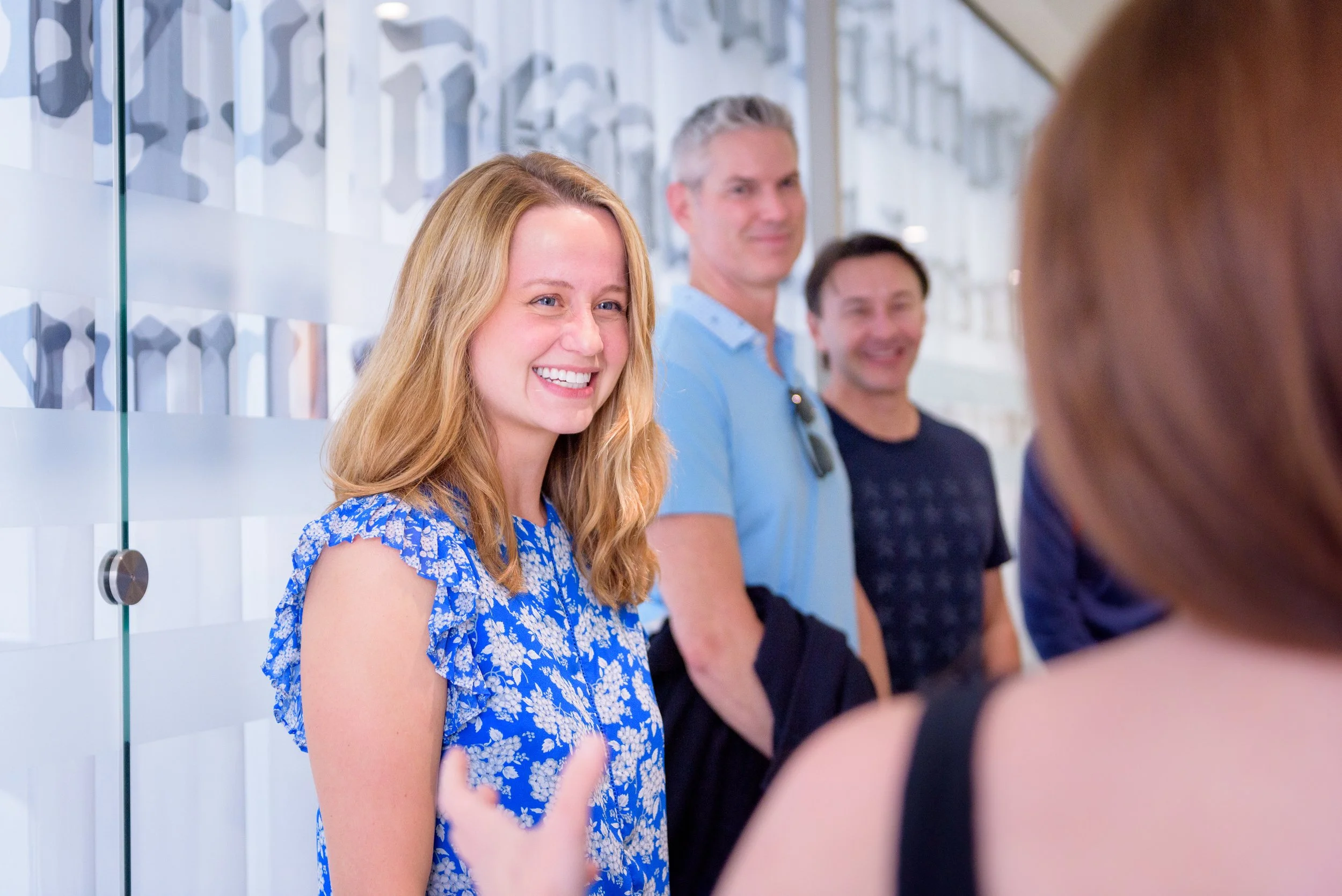 Four people standing in line, talking, with a woman in a blue floral dress smiling, and a man with gray hair and a blue shirt smiling, in a bright indoor setting.