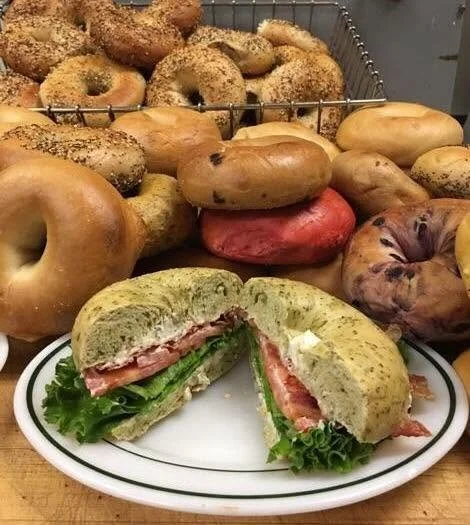 A bagel sandwich with lettuce, tomato, and cream cheese on a white plate, with various bagels in the background.