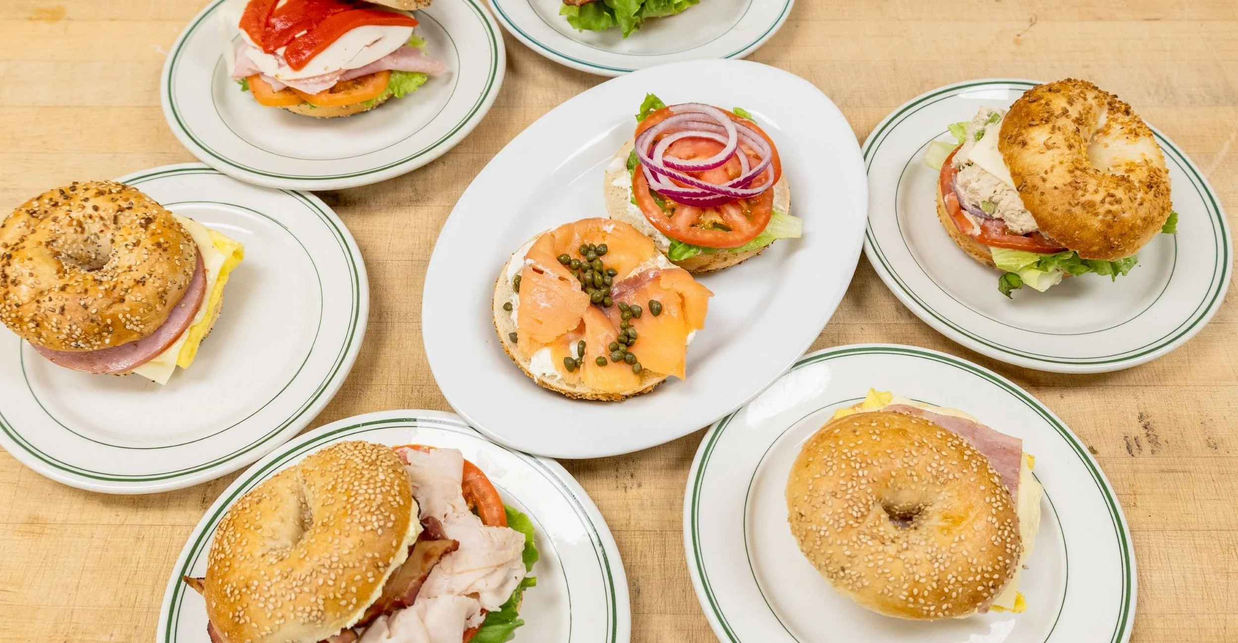 Assorted sandwiches on white plates with green stripes, some with sesame seed buns, filled with various meats, cheeses, lettuce, tomatoes, onions, and other toppings, arranged on a wooden table.