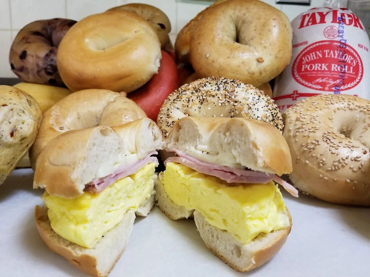 Breakfast sandwiches with scrambled eggs, ham, and bagels, surrounded by various bagels and a pack of Taylor pork roll.