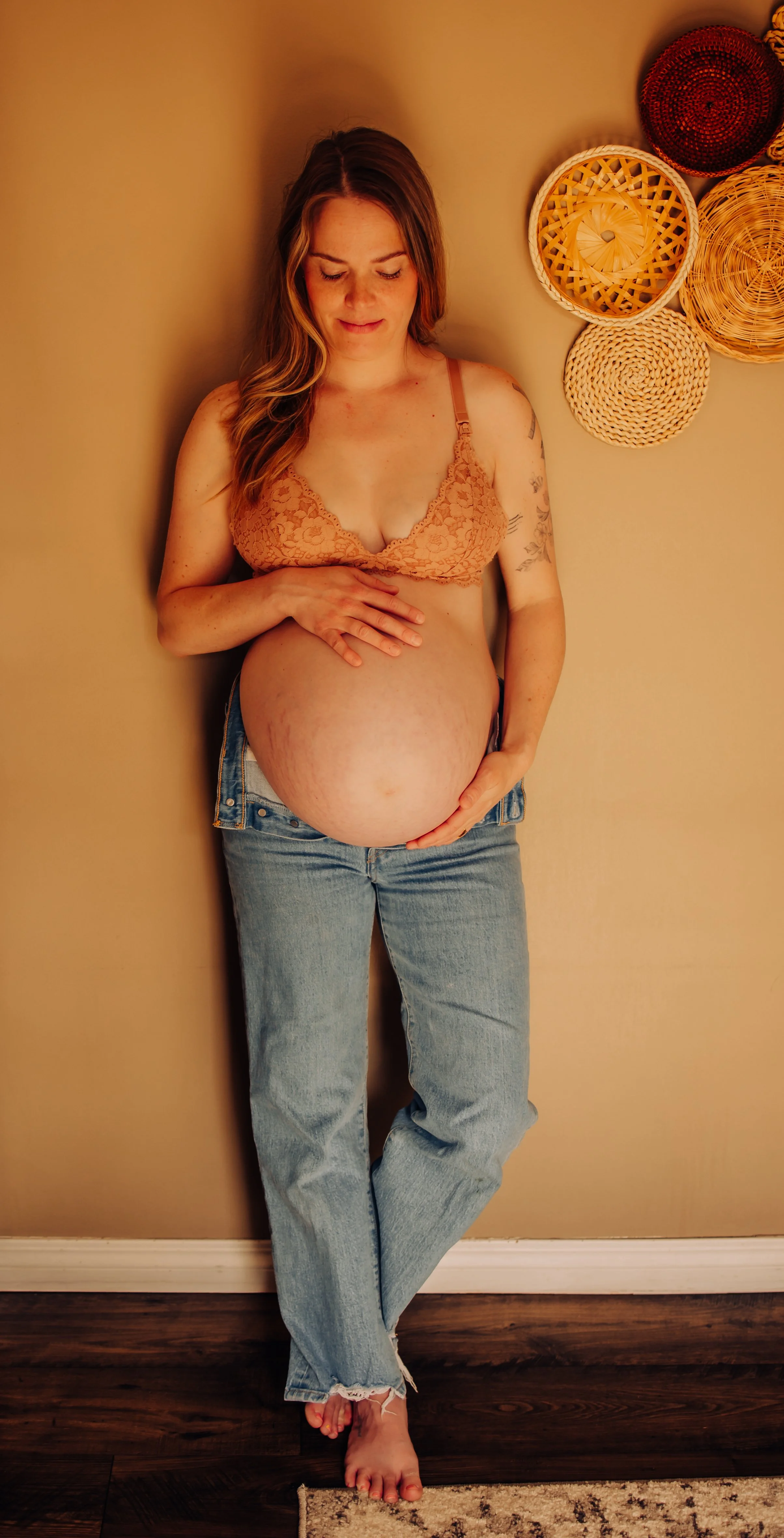 A pregnant woman stands indoors against a beige wall decorated with woven baskets, with her eyes closed and a gentle smile, wearing a peach lace bra and light blue jeans.