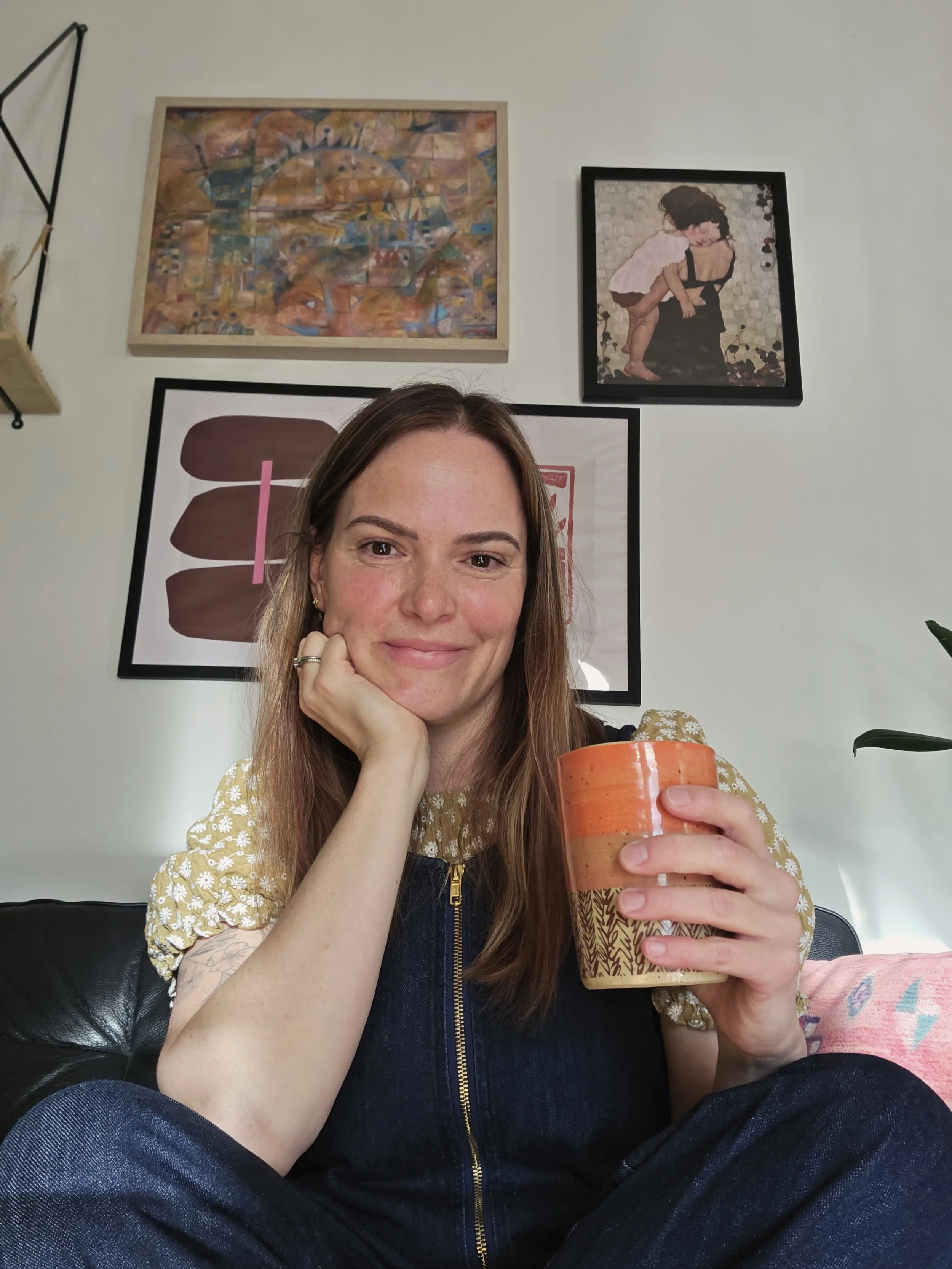 A woman with long brown hair and a yellow floral top is sitting on a black sofa, smiling and holding a large colorful coffee mug, with her chin resting on her hand in a cozy room decorated with framed artwork and a plant.