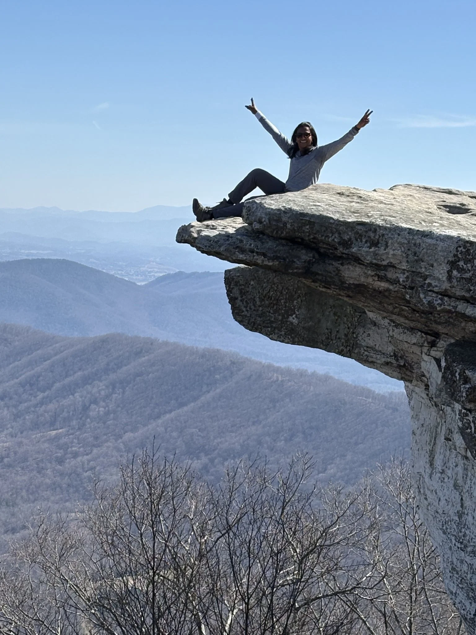 Hiker sitting on a rocky mountain overlook with arms raised above a forested mountain landscape.