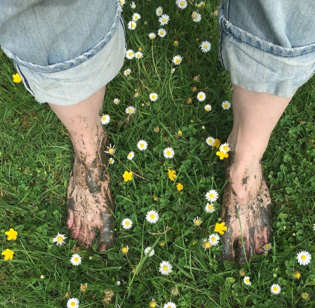 Mud-covered bare feet and rolled-up jeans standing in a grassy field dotted with daisies and yellow flowers