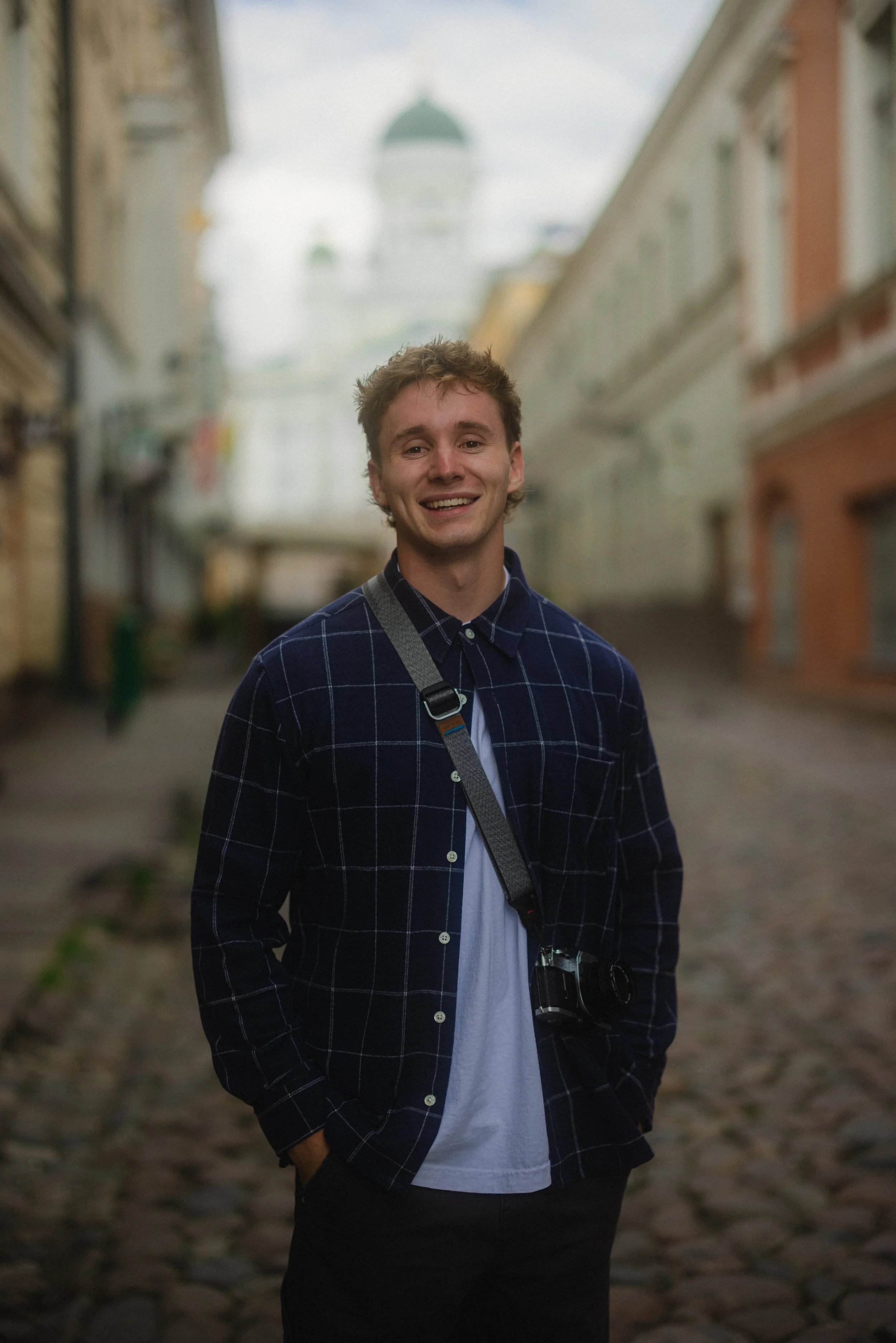 A young man with curly hair smiling, standing on a cobblestone street with blurred buildings in the background, and a church dome in the distance.