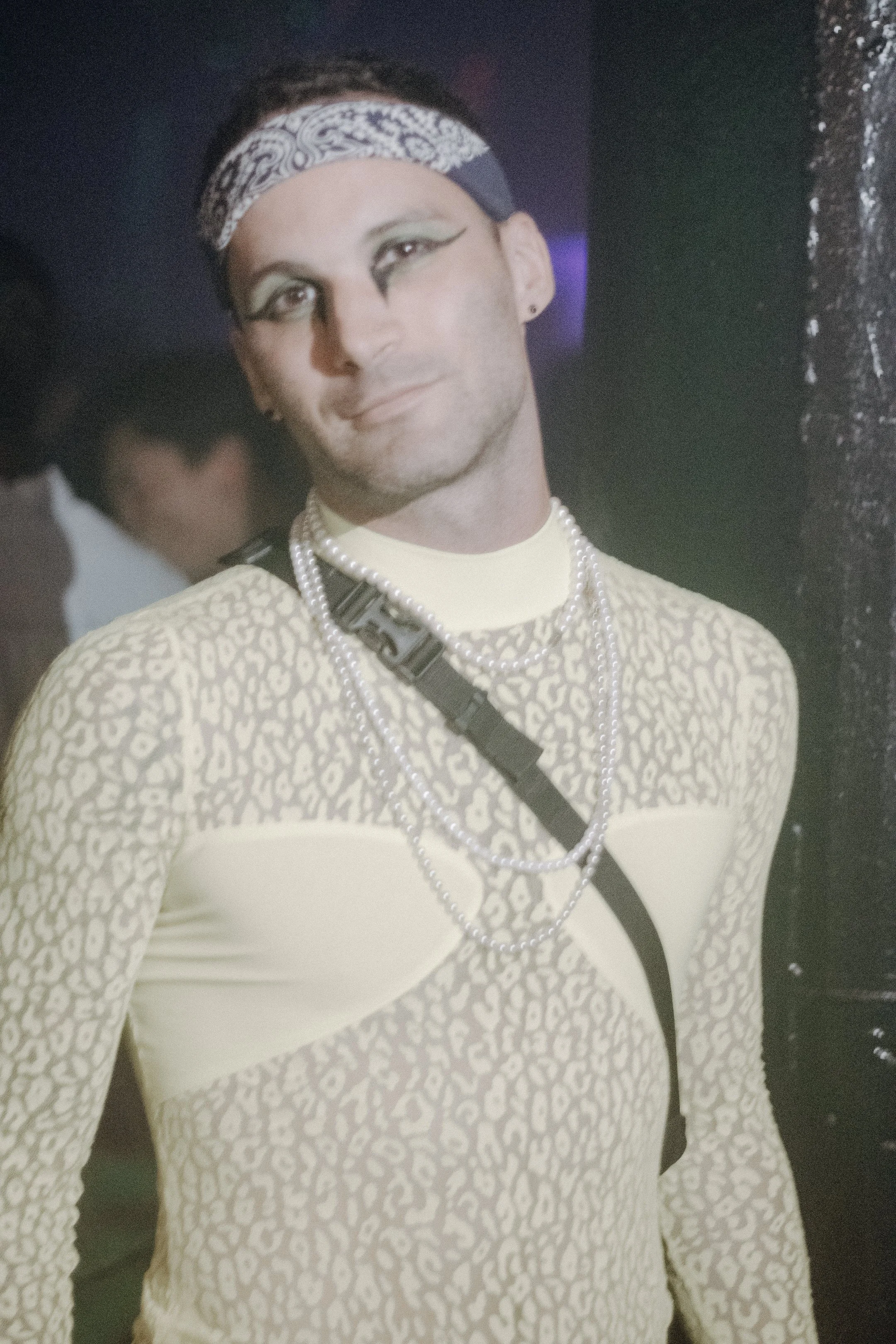Performer in a bandana, patterned shirt and layered necklaces, meeting the camera