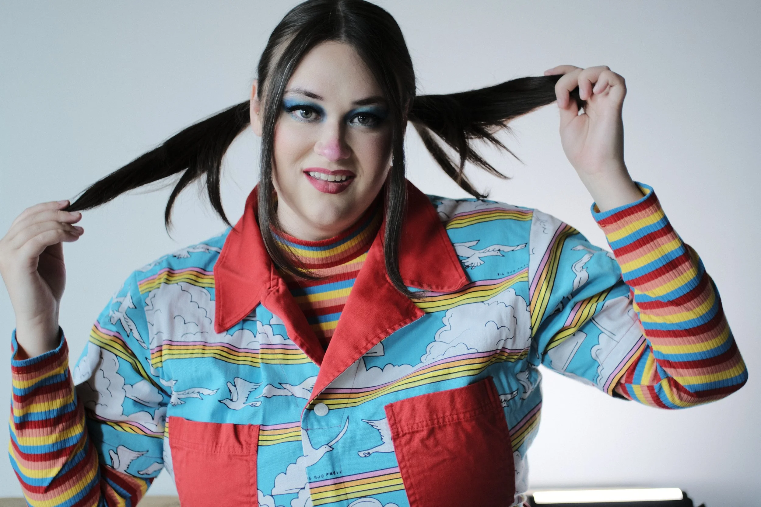 Hands pull the pigtails wide above a rainbow-striped collar.
