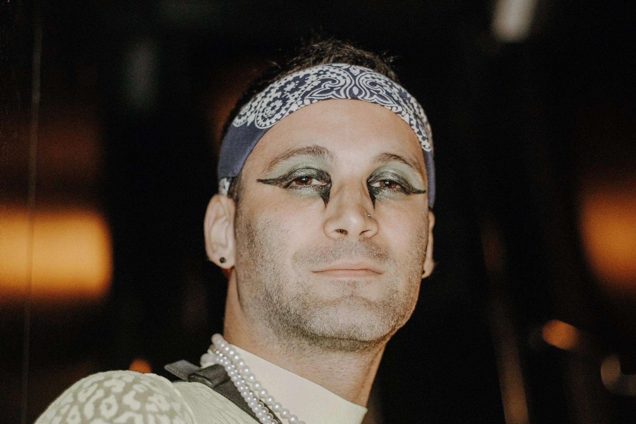 Performer in bandana and dramatic eye makeup, staring into the lens under warm club light