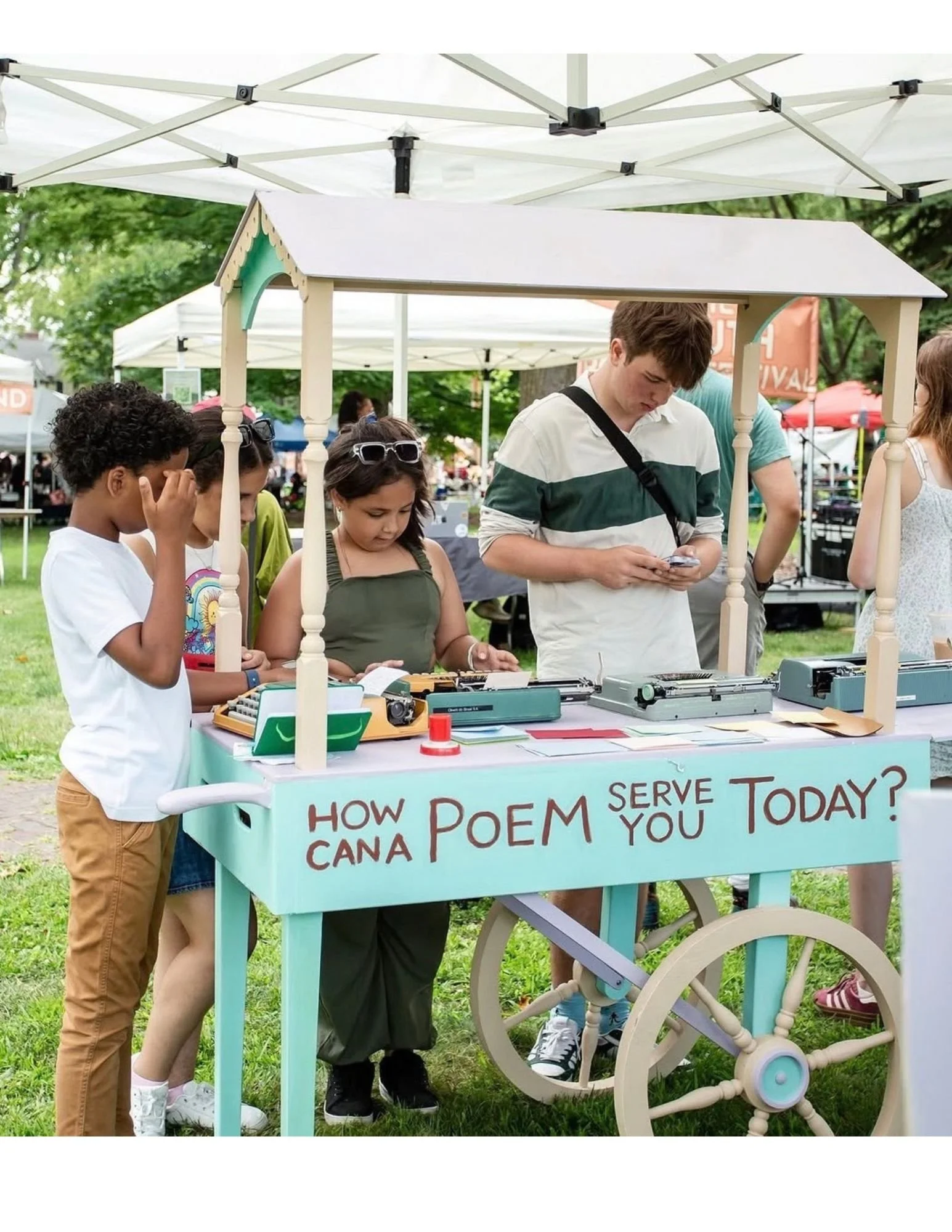 Children and a young man gathered around a small vintage cart at an outdoor event, with a sign asking, "How do I poem serve today?" on the cart. Tents and trees are visible in the background.