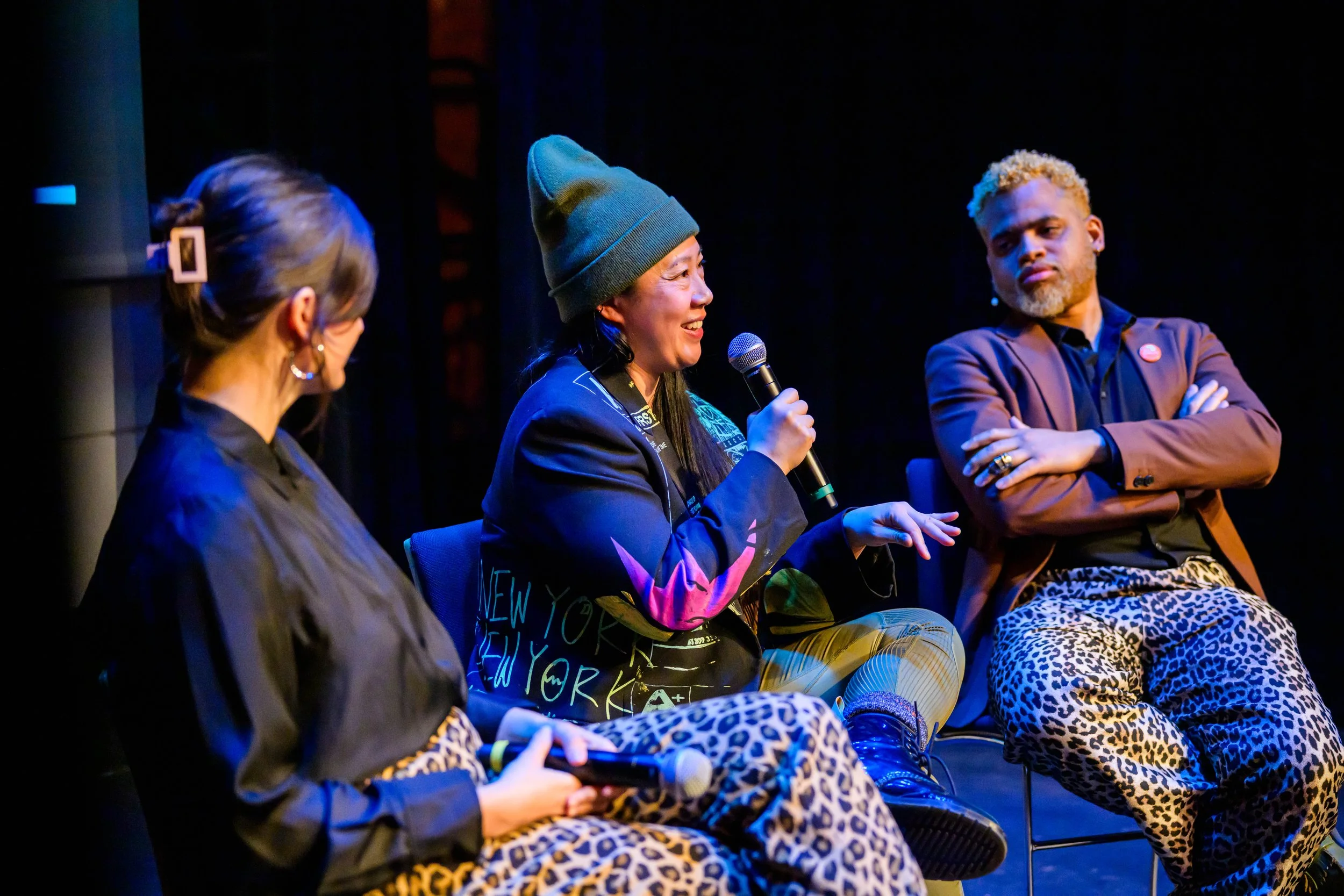 Three women are seated on stage participating in a panel discussion. The woman in the middle, with long dark hair and wearing a beanie, is speaking into a microphone. The woman on the left has gray hair tied back and is looking down. The woman on the right has short blonde hair and is wearing leopard print pants, listening with arms crossed. The setting is dimly lit with a dark background.