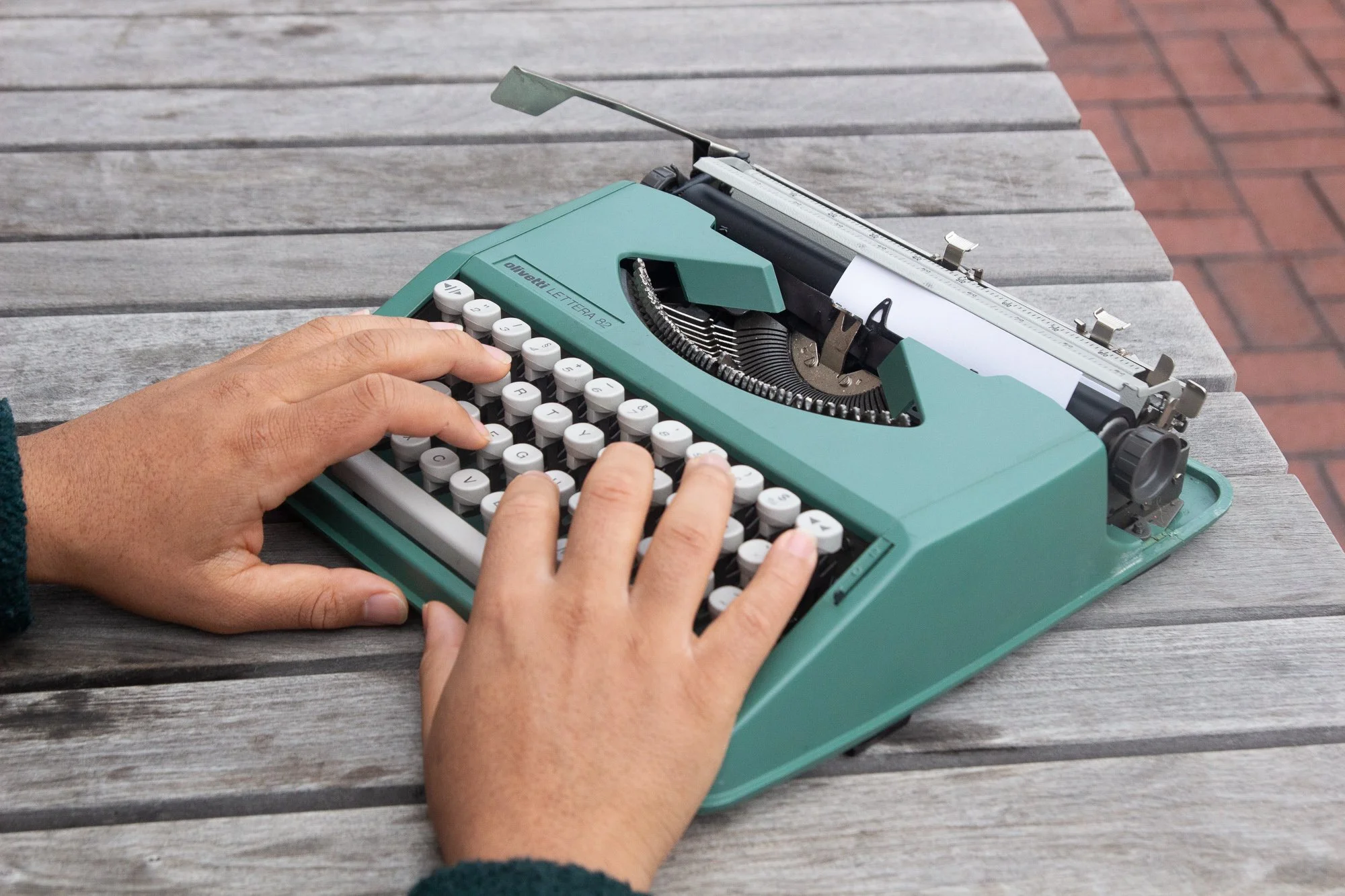 Person typing on a vintage turquoise typewriter placed on a wooden table outdoors.