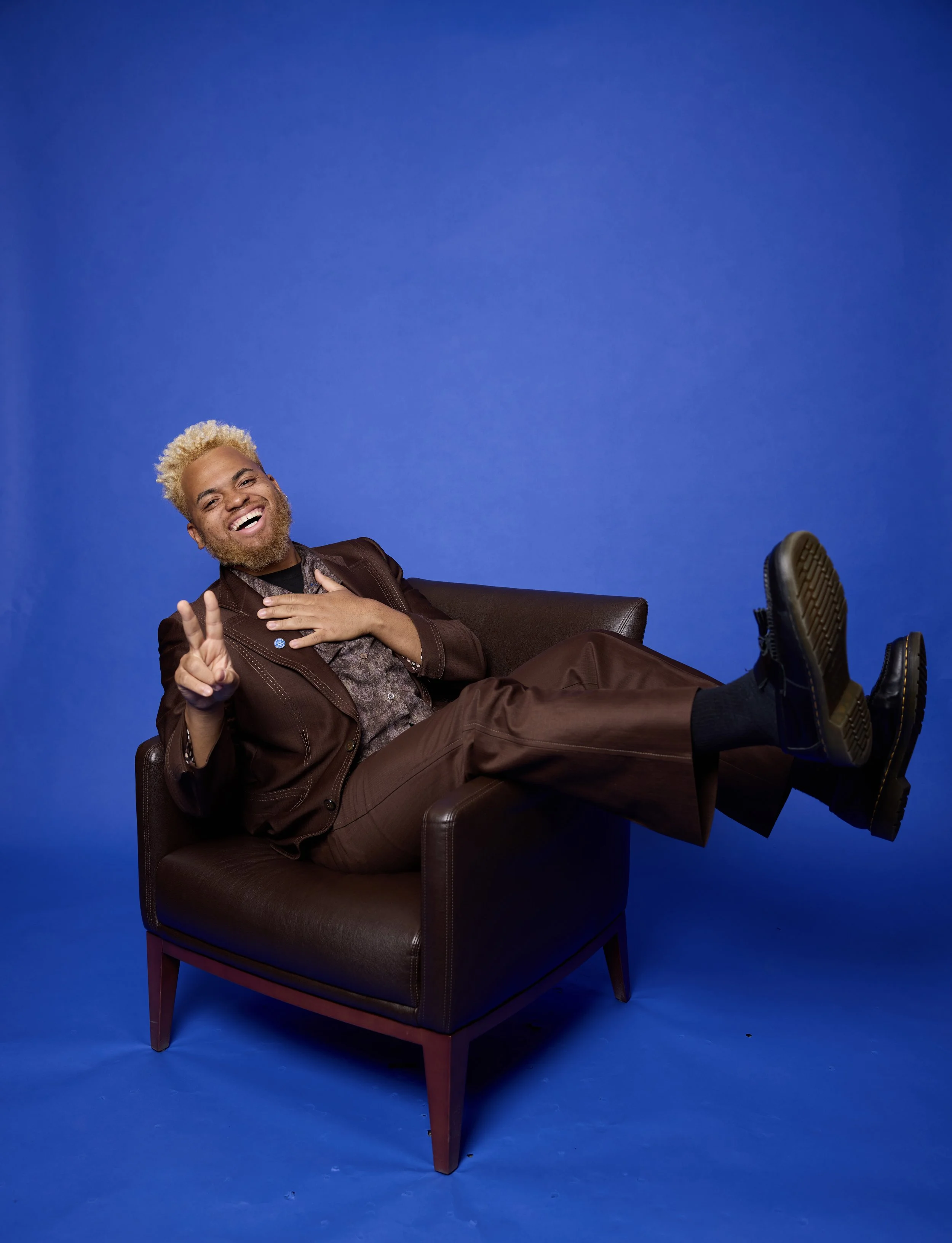 A smiling man with blond curly hair and a beard, dressed in a brown suit and patterned shirt, sitting in a leather chair against a solid blue background, with his feet propped up on the chair and making a peace sign with his fingers.