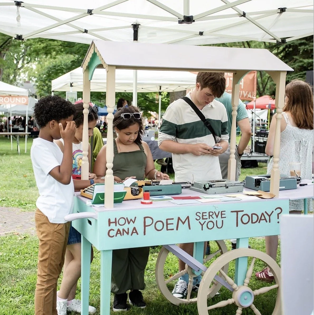Children and a young man at a poetry vending machine booth outdoors during a festival. The booth has a sign that reads, 'How can poem serve you today?'