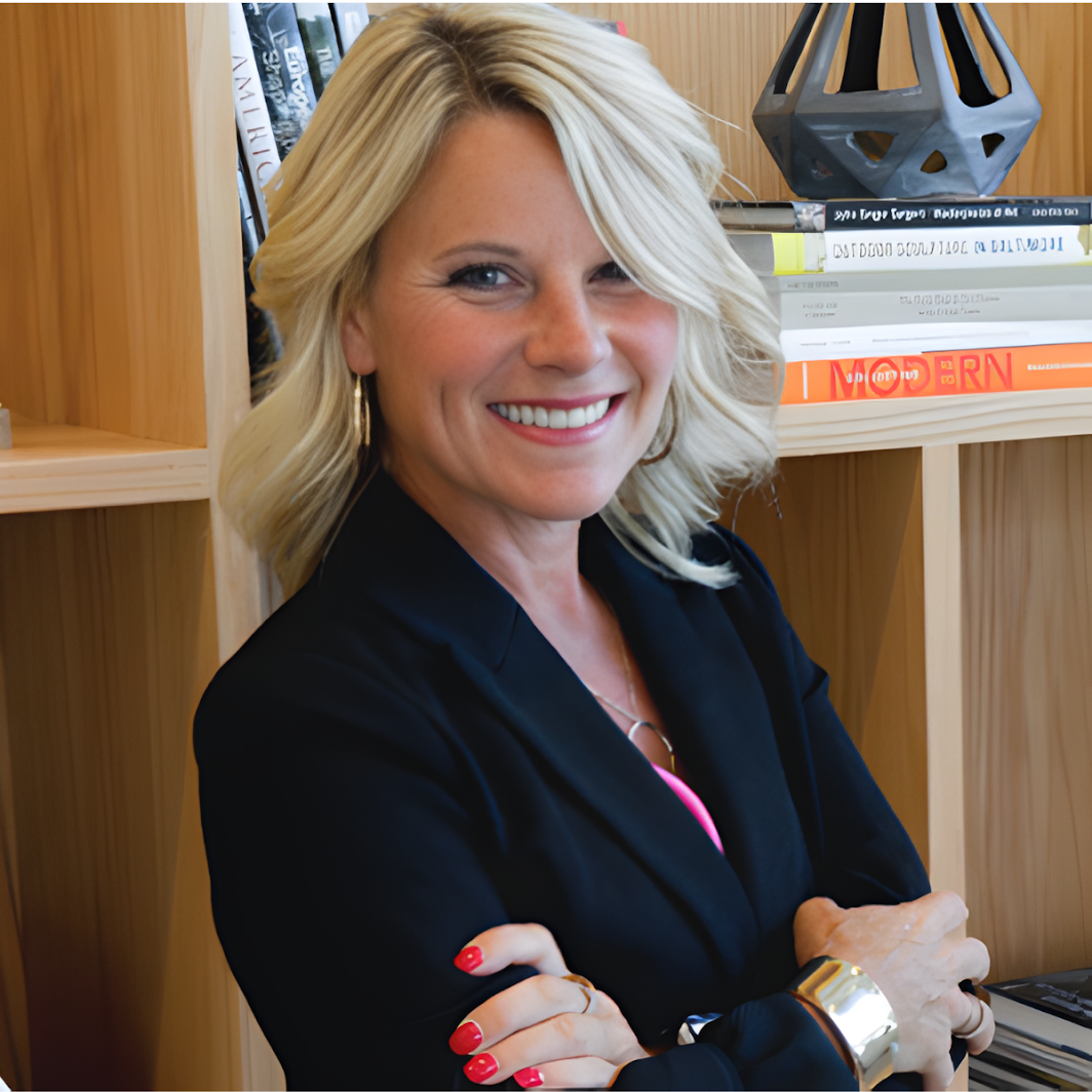 A smiling blonde woman wearing a black blazer, standing in front of wooden shelves filled with books and decorative items.