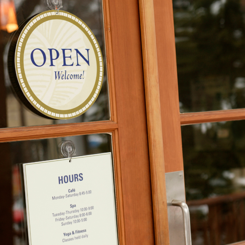 Wooden door window with an "Open" sign that says "Welcome!" and a white hours notice listing cafe, spa, and yoga & fitness hours.