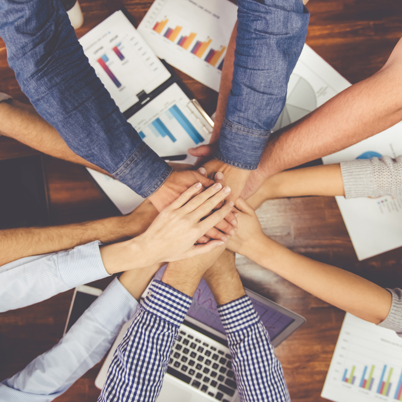 Group of diverse people stacking hands in a teamwork gesture on a wooden table with charts and a laptop.