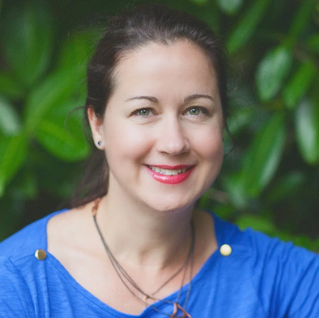 A woman with dark brown hair and blue eyes smiling, wearing a blue top and layered necklaces, with green leaves in the background.