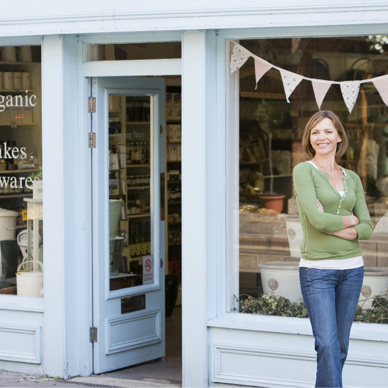 A woman standing outside a light blue shop with large windows decorated with bunting, smiling with arms crossed.