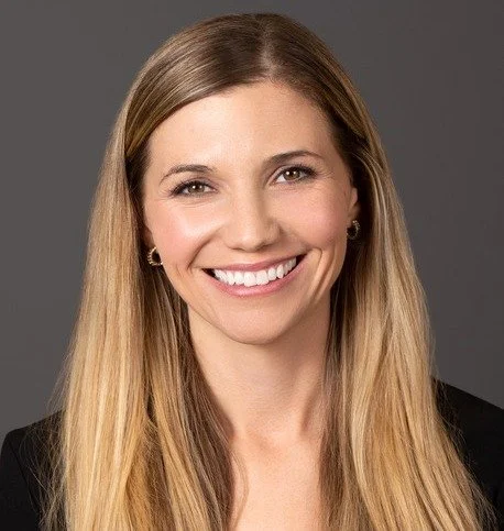 A woman with long blonde hair smiling against a gray background.