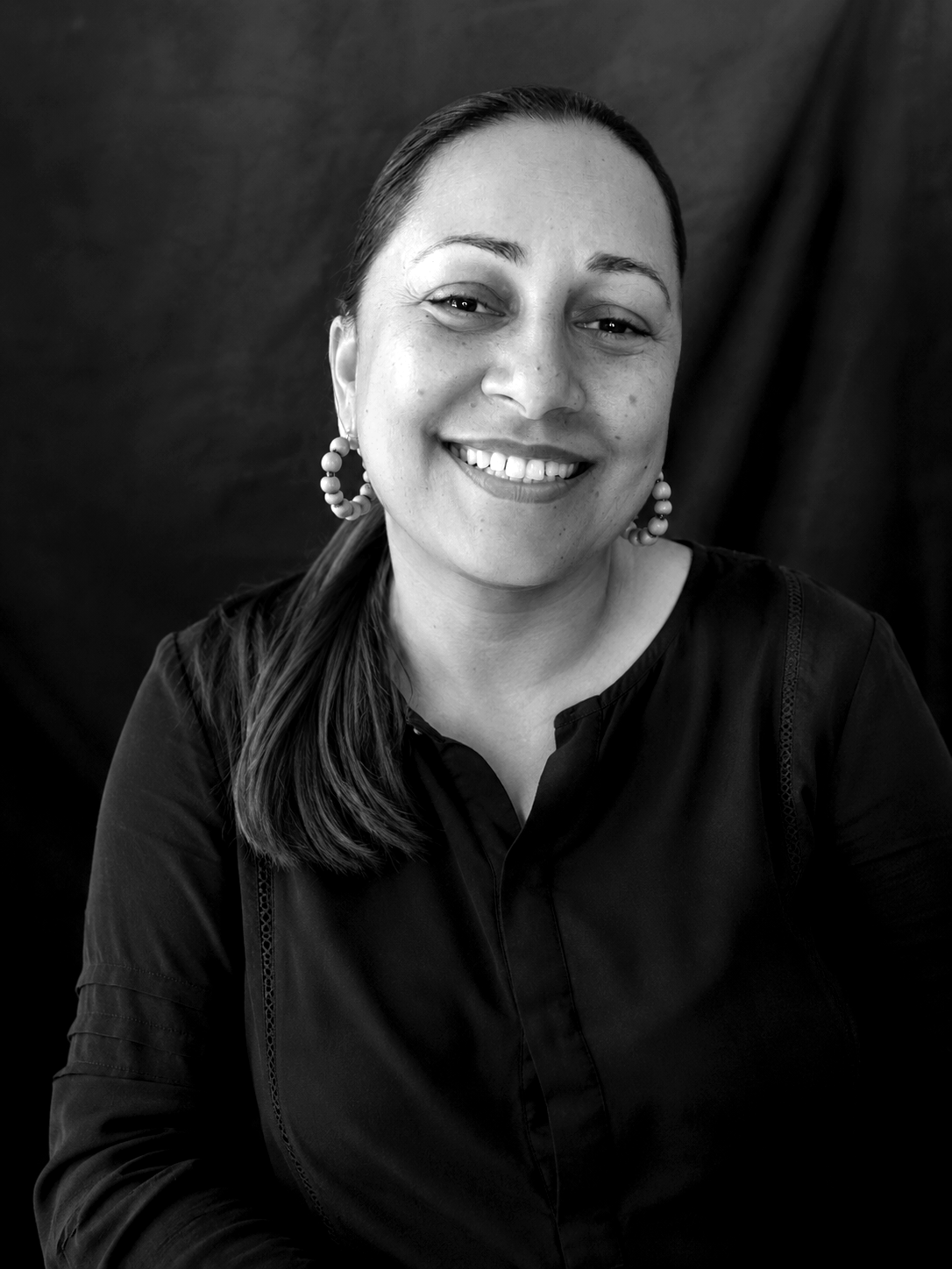 Black and white portrait of a smiling woman wearing earrings and a dark top, posing against a dark background.